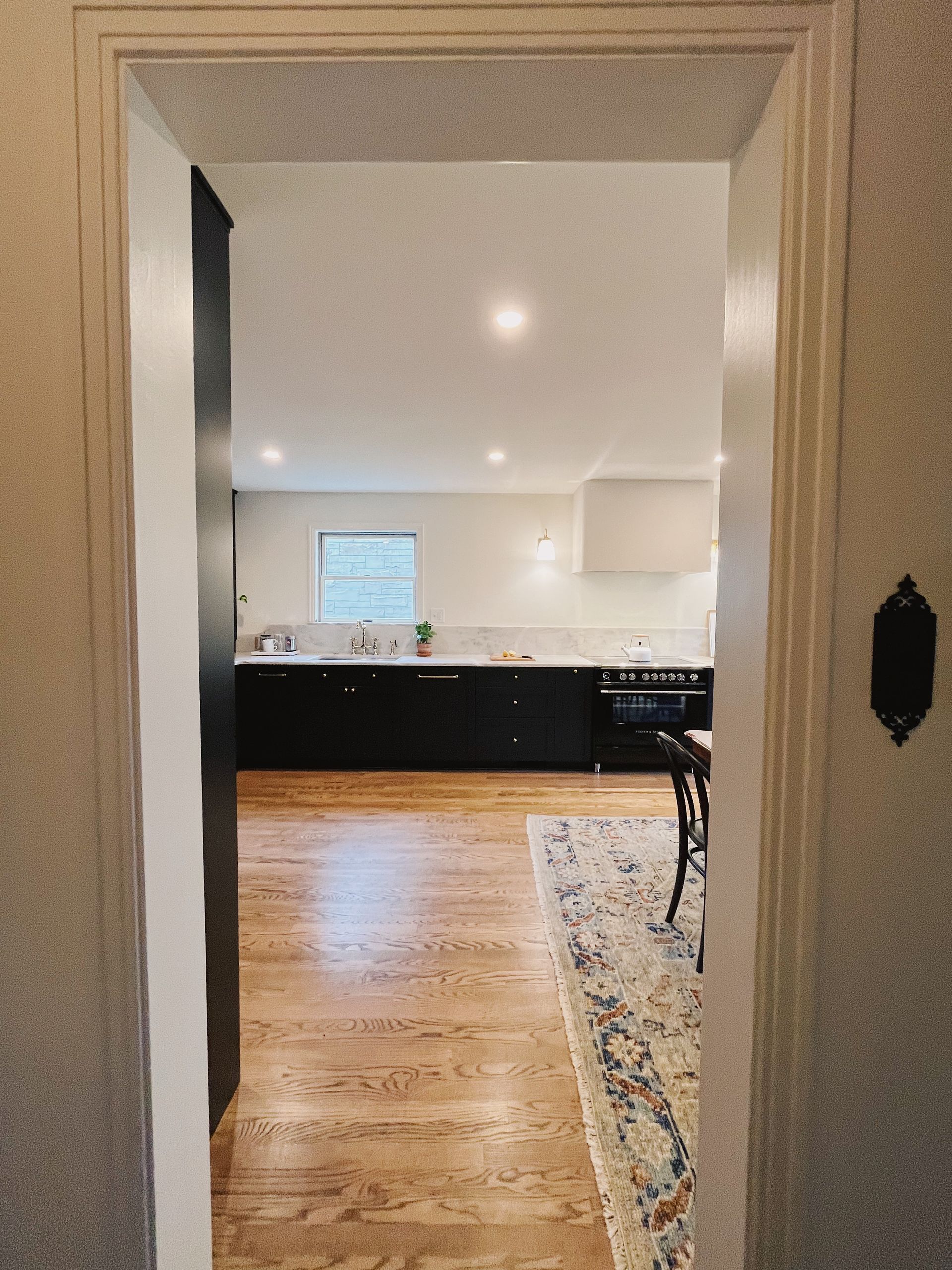 View through a doorway into a bright kitchen with dark cabinets, white walls, wood floors, and a rug.
