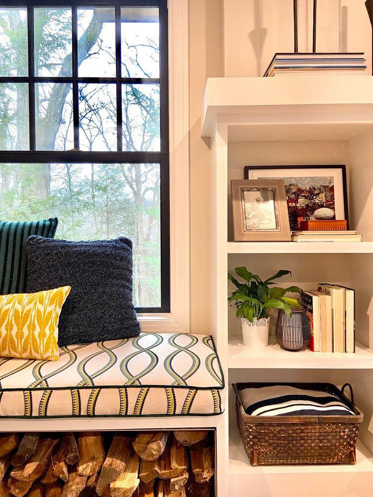 Cozy window seat with patterned cushions beside a built-in bookshelf and a stack of firewood underneath.