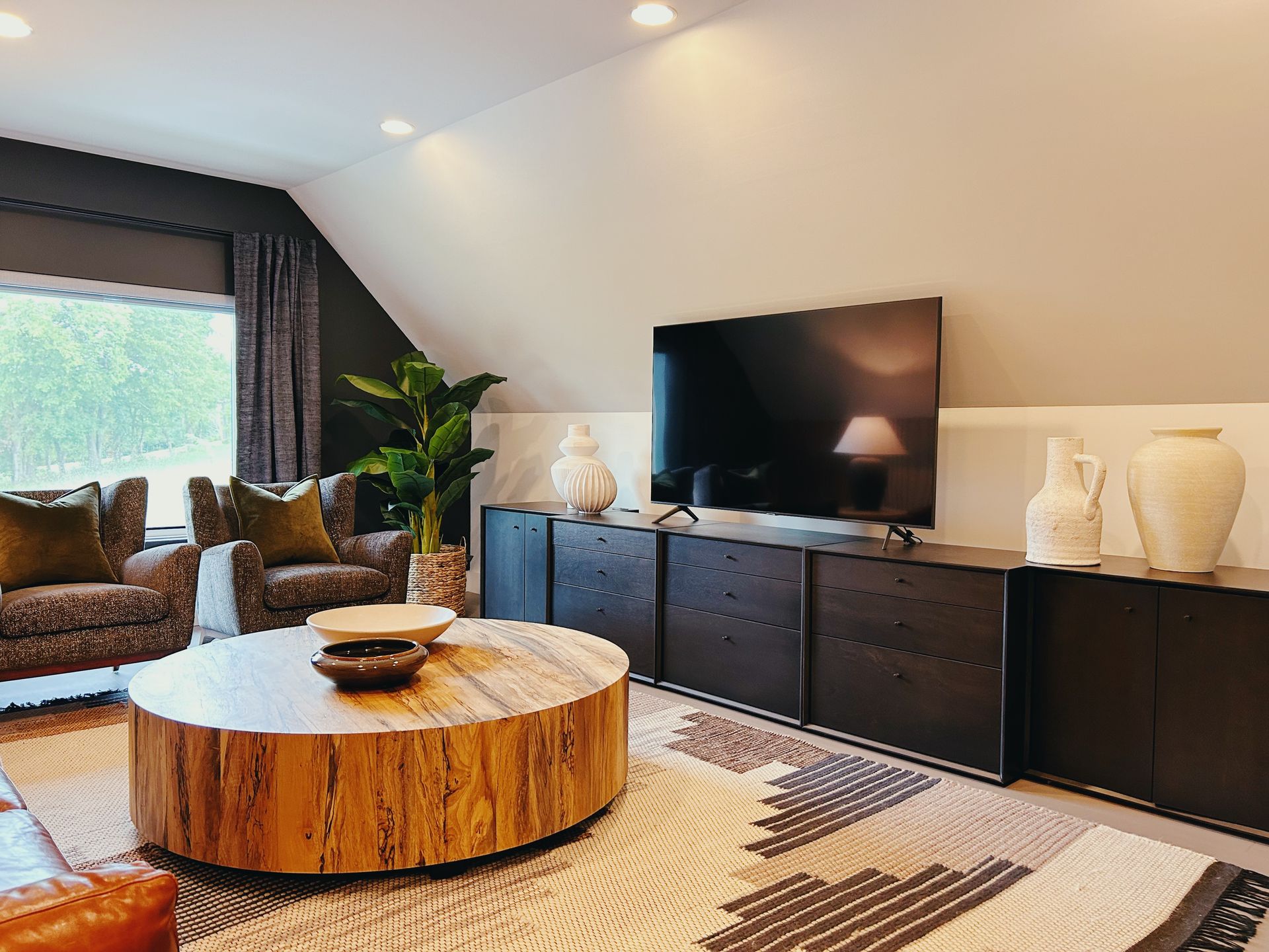 Modern living room with a TV console, round wood coffee table, sofa, plants, and a large window