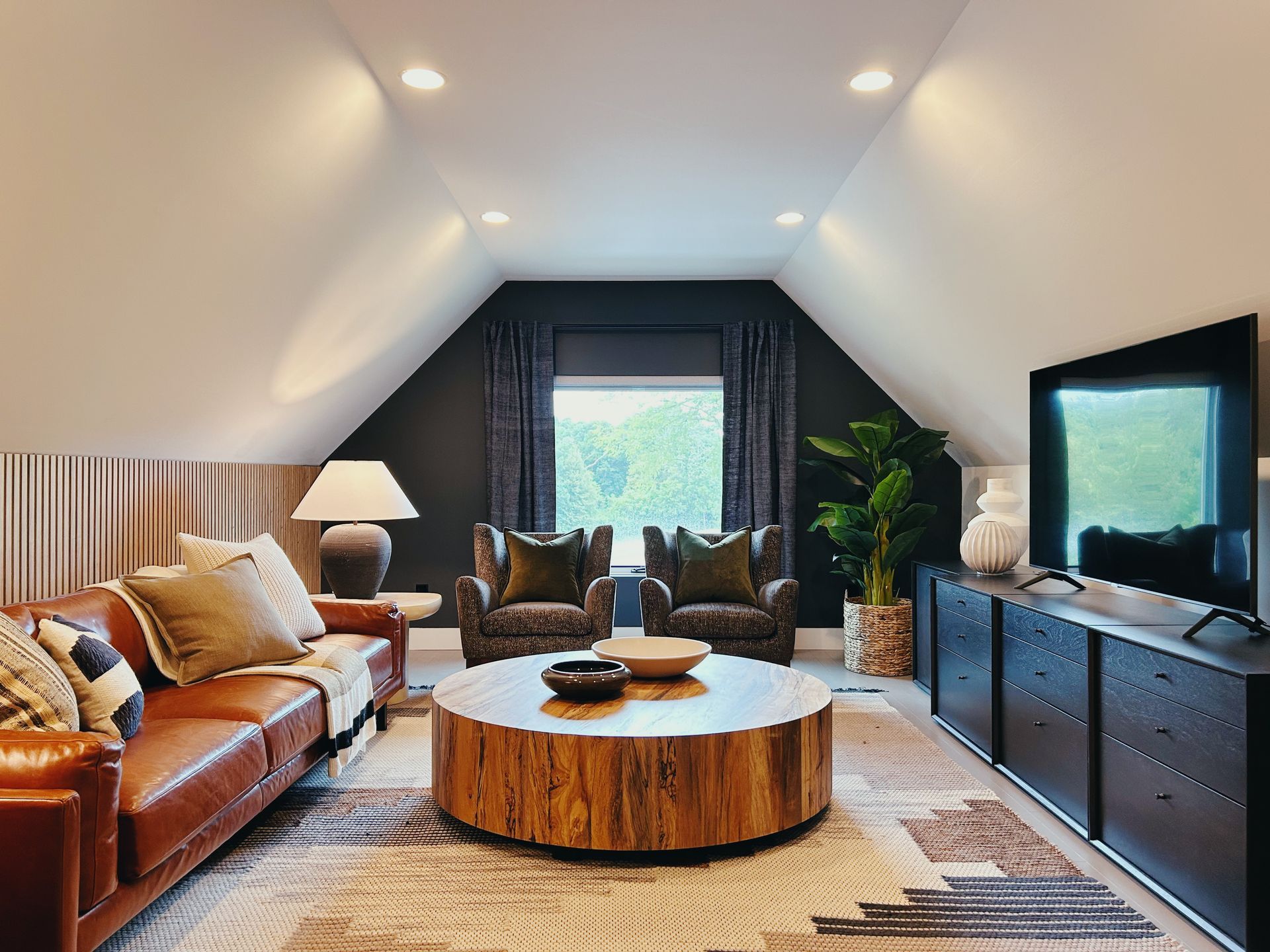 Bright attic living room with leather sofa, round wood coffee table, armchairs, and a TV console.