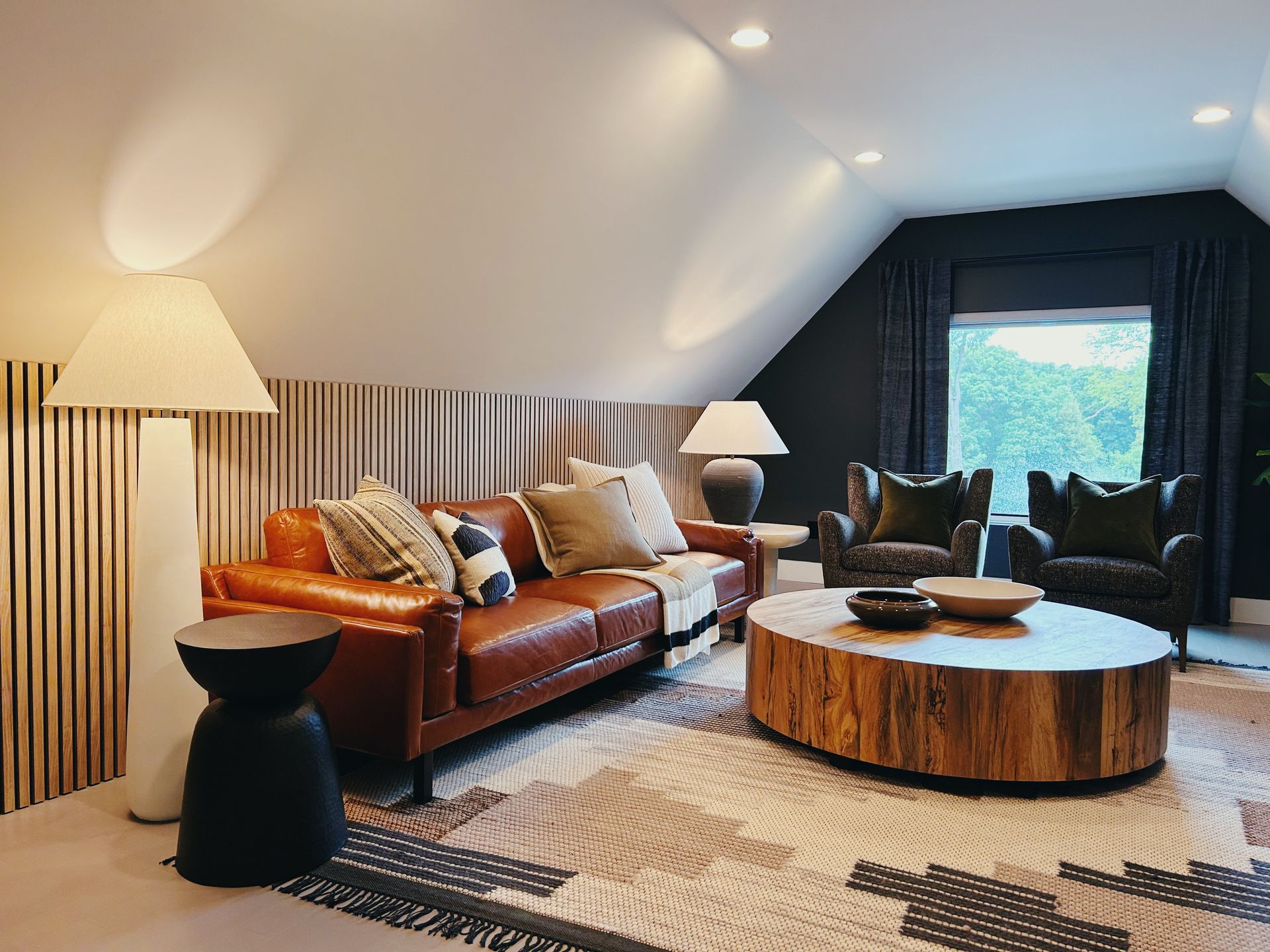 Cozy attic living room with brown leather sofas, round wood coffee table, floor lamps, and a large window view