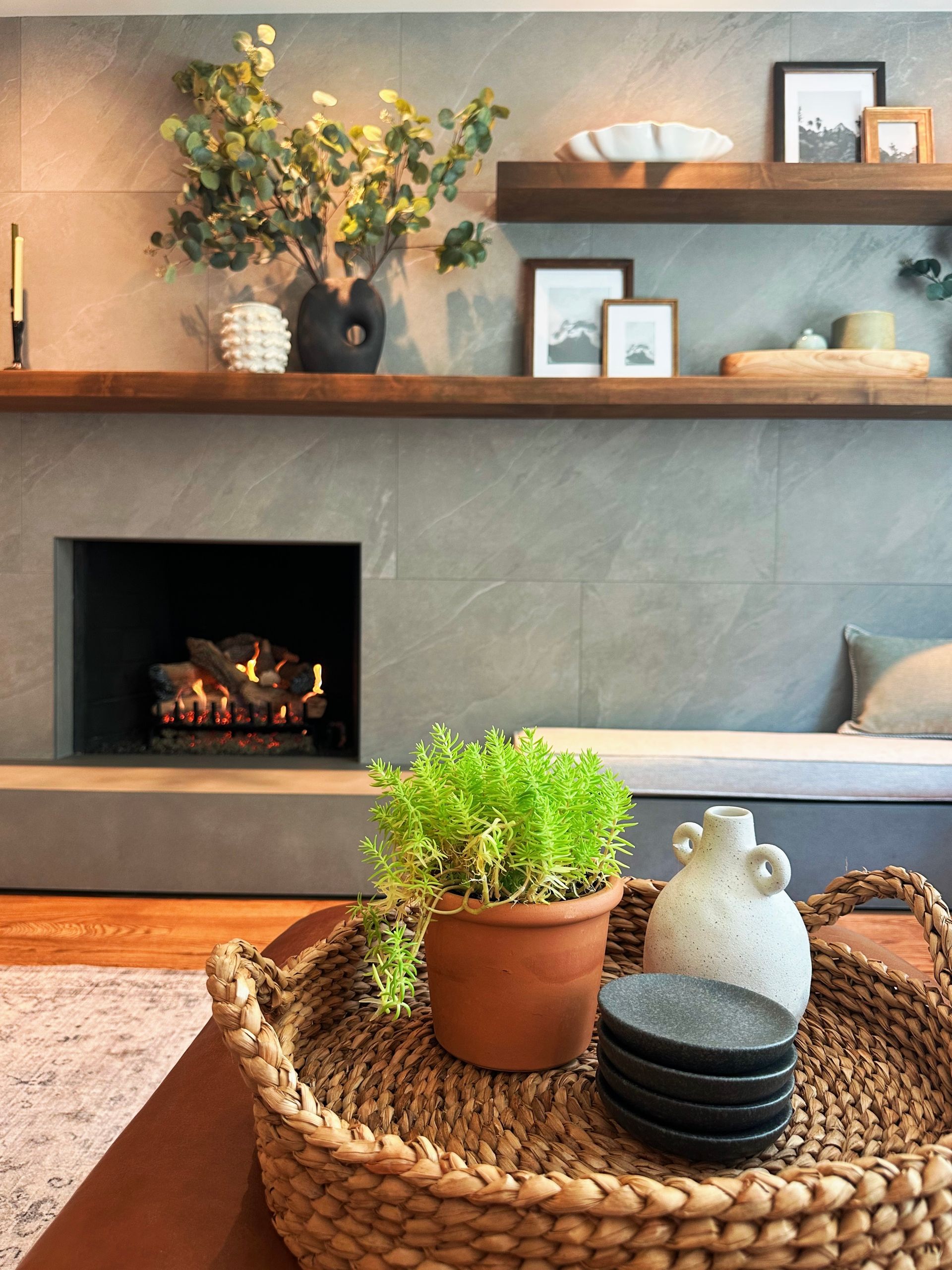 Cozy living room with a lit fireplace, shelf decor, and a woven tray holding a potted plant and ceramics