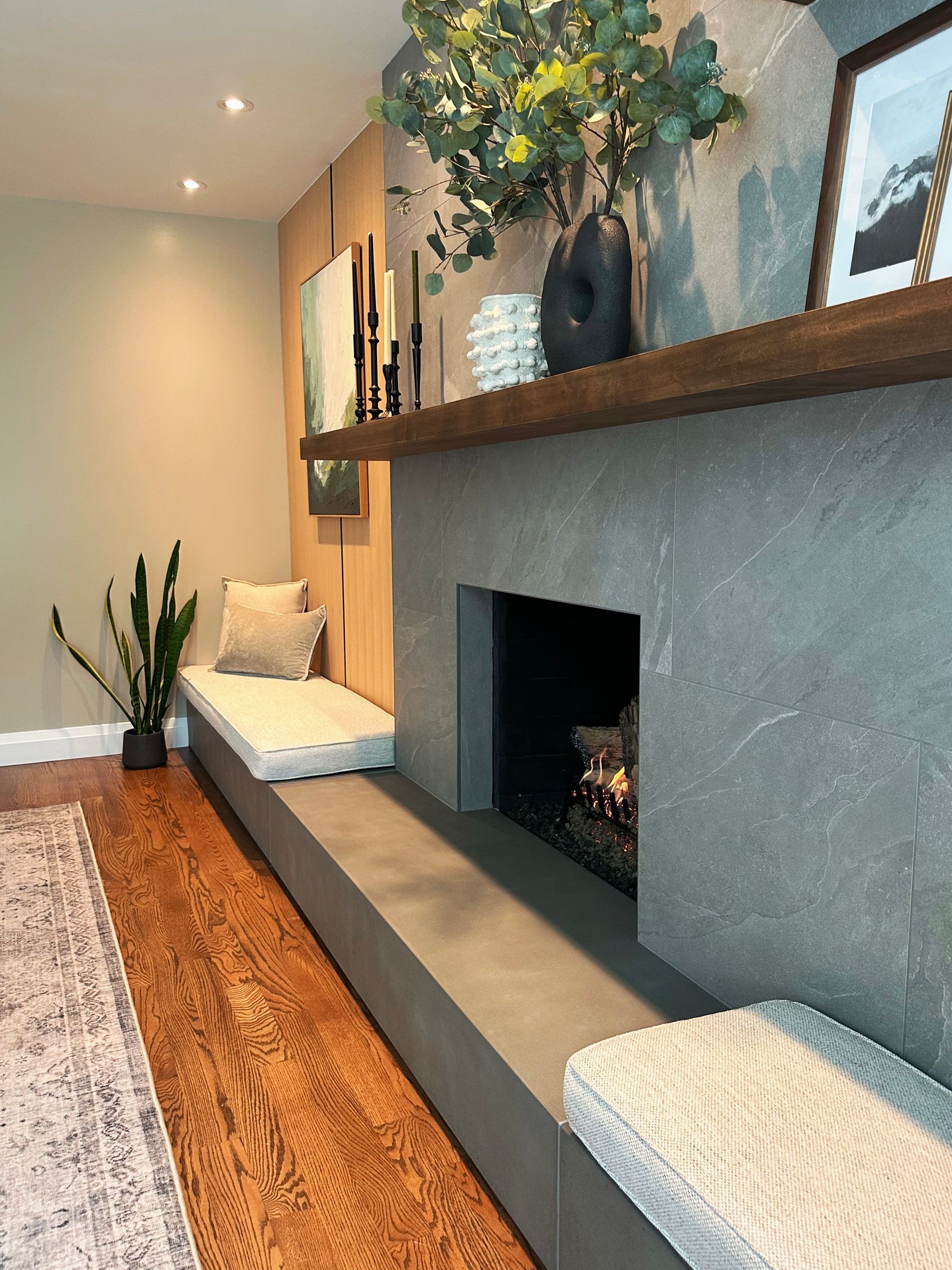 Modern living room with a gray fireplace, wooden mantel, plants, and a patterned rug