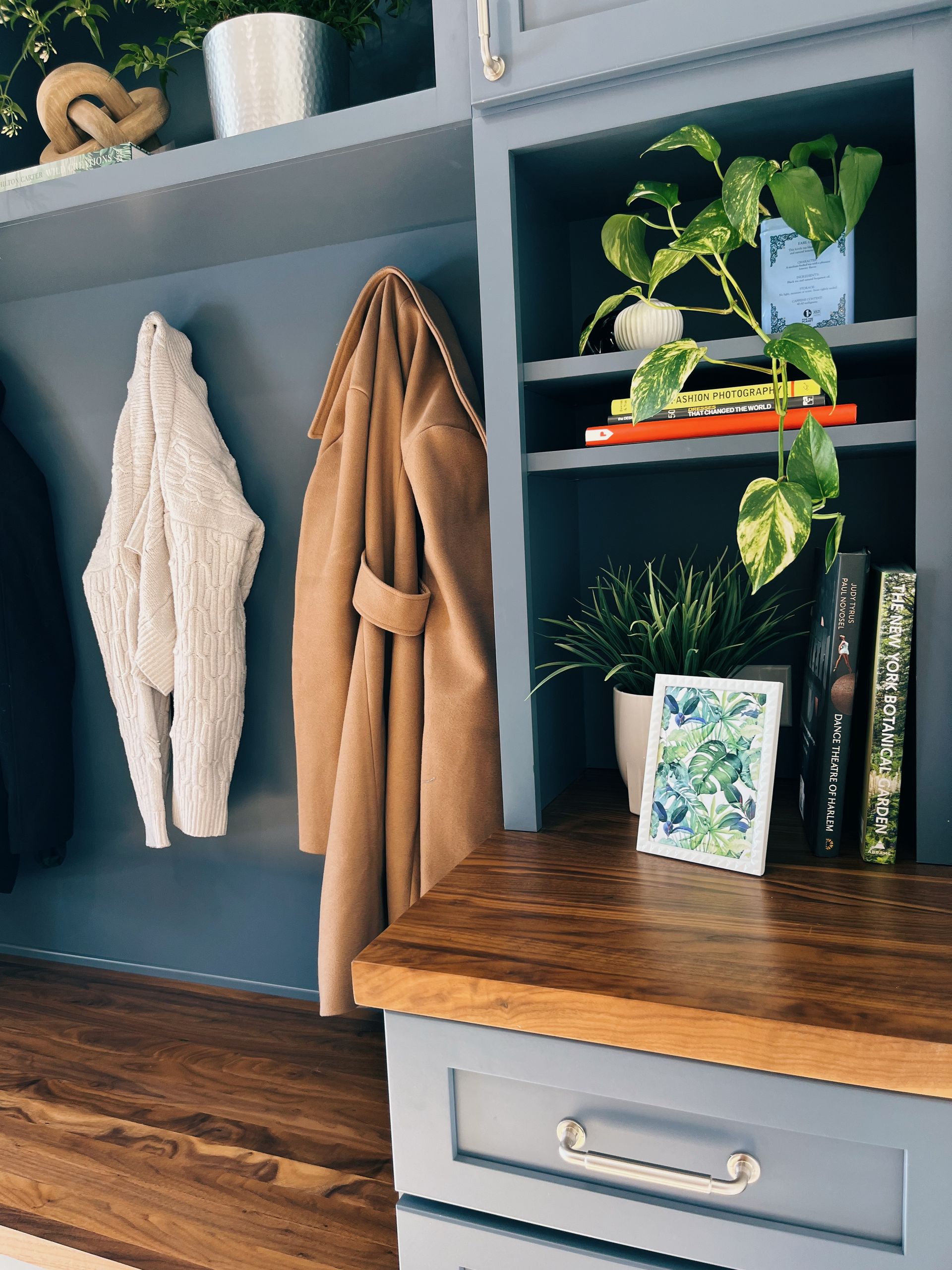 Coat hooks with a white knit and tan robe beside a wood bench, shelf, plant, and books in a mudroom.
