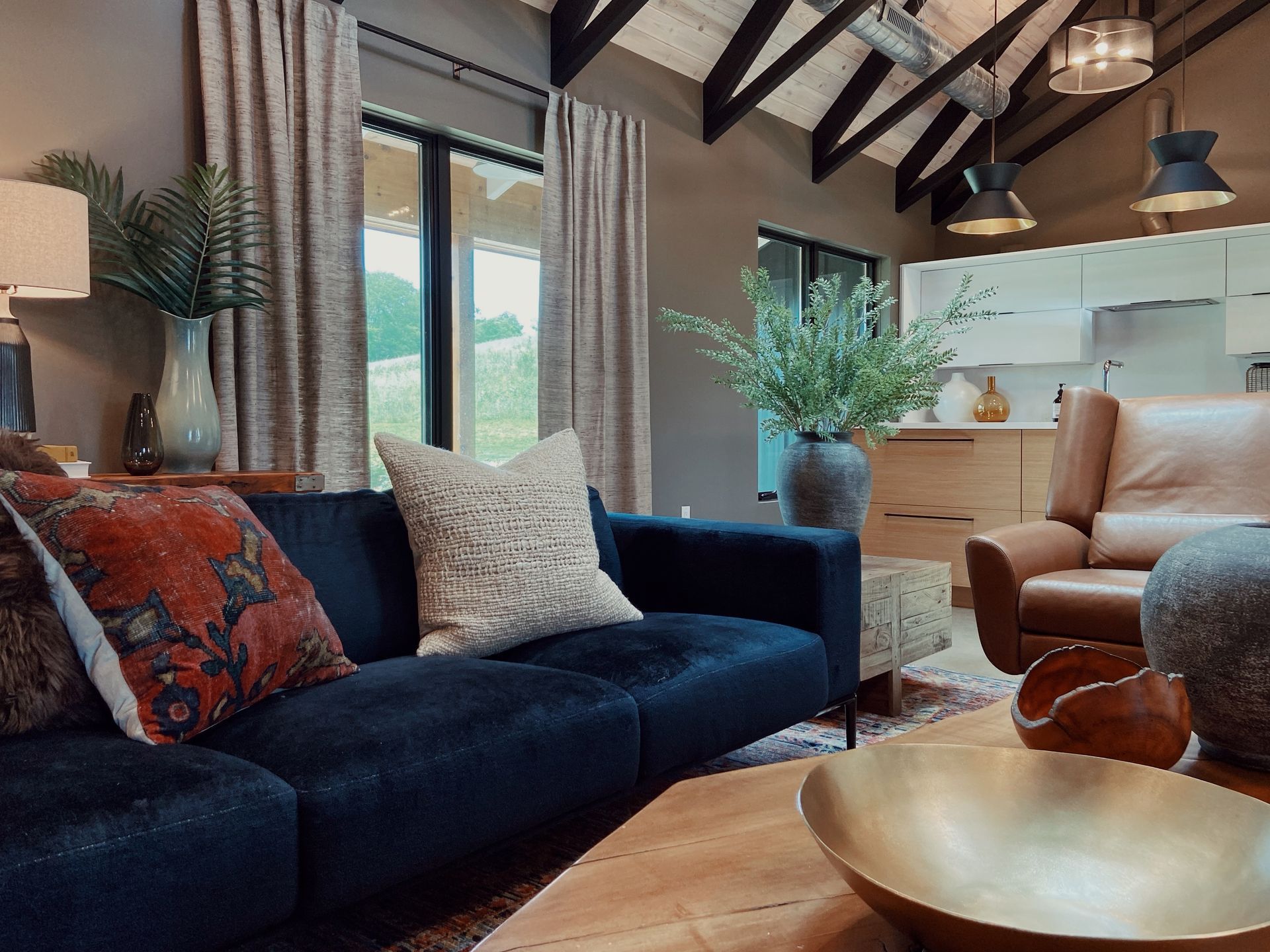 Cozy living room with dark sofa, tan chair, wood beams, and mountain view through large windows