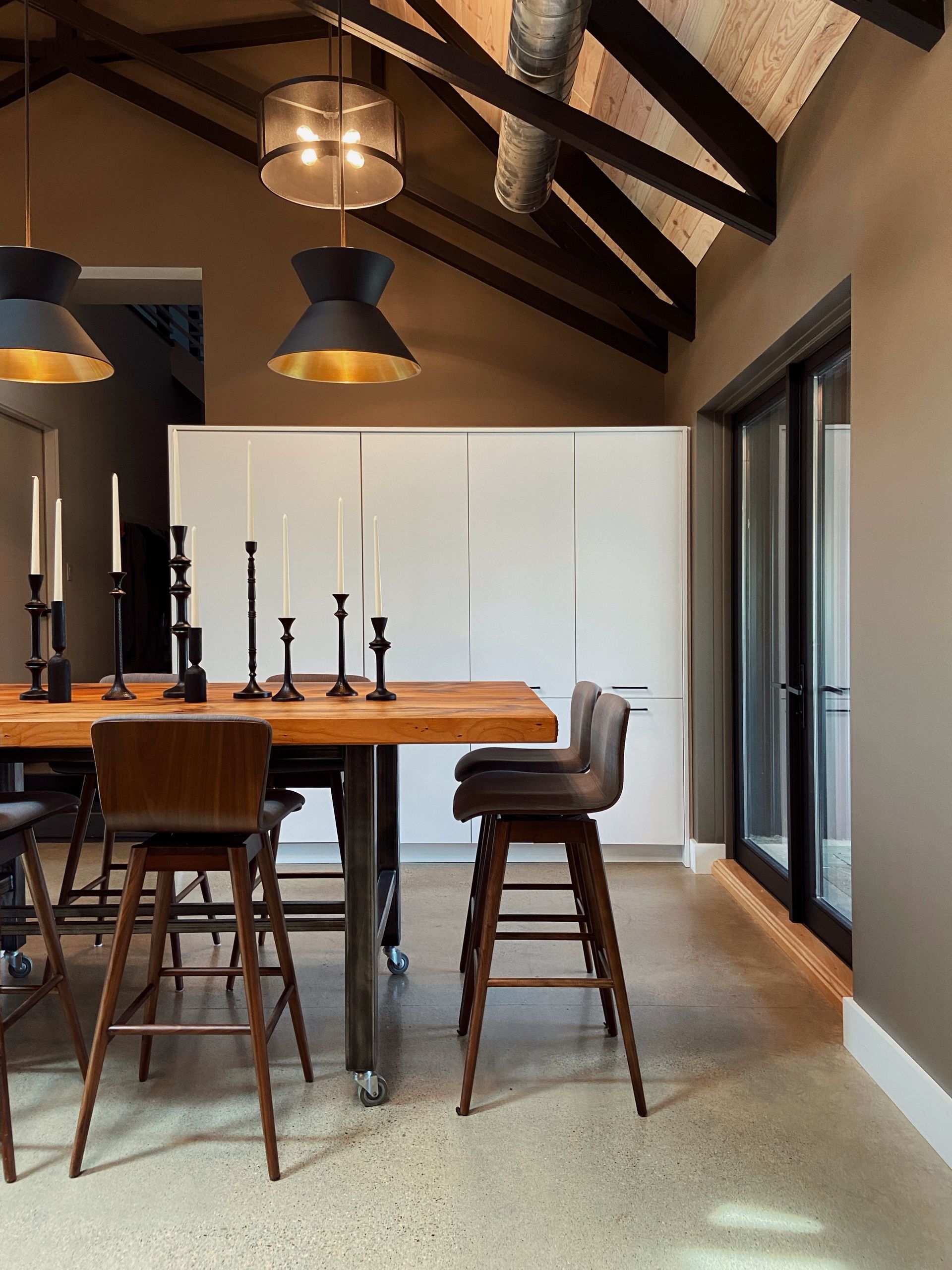 Modern dining room with wooden table, bar stools, pendant lights, and vaulted ceiling beams