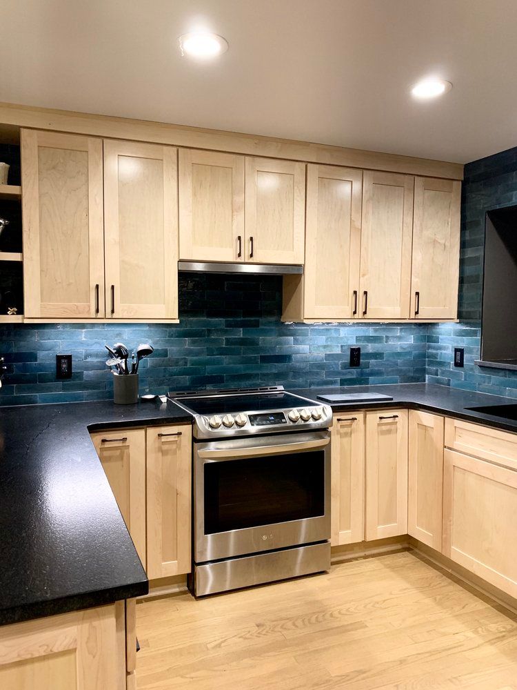 Modern kitchen with light wood cabinets, blue tile backsplash, stainless steel stove, and black countertops.