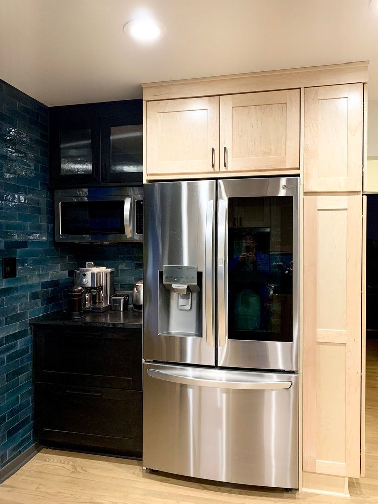 Modern kitchen with stainless steel refrigerator, light wood cabinets, dark counters, and teal tile backsplash