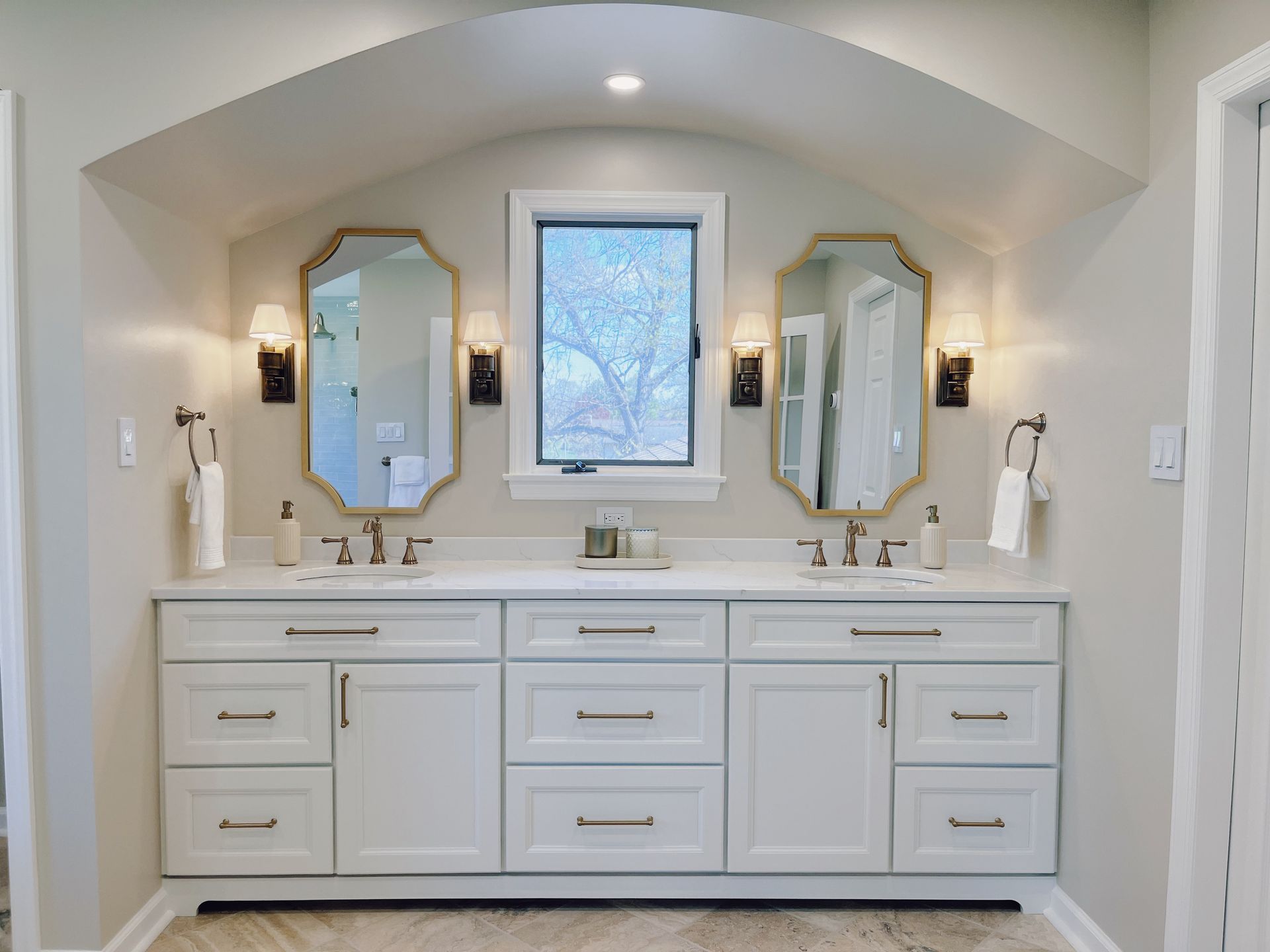 Bright bathroom vanity with double sinks, mirrors, sconces, and a central window in a light neutral room