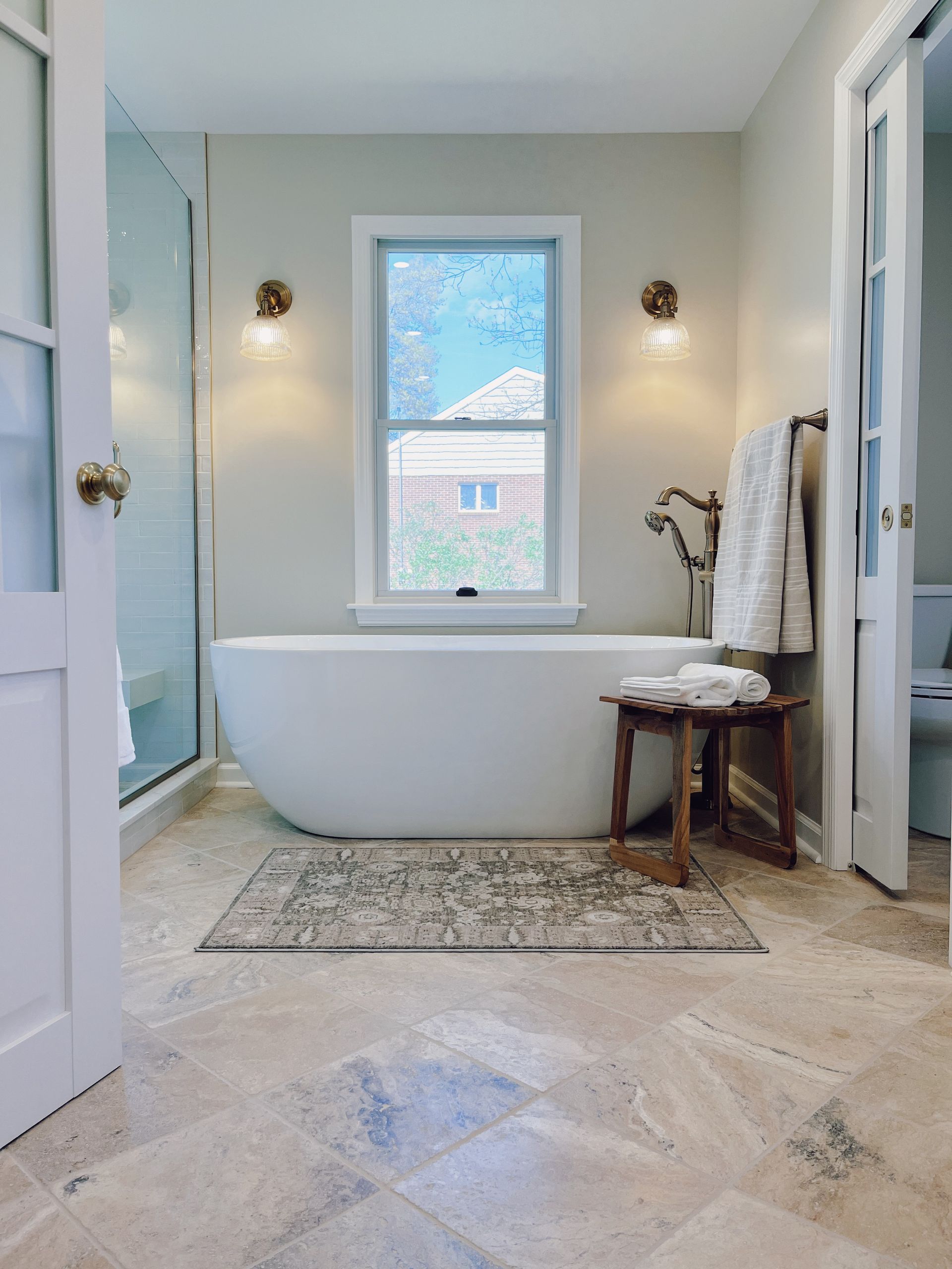 Bright bathroom with freestanding tub, window, wall sconces, and patterned rug
