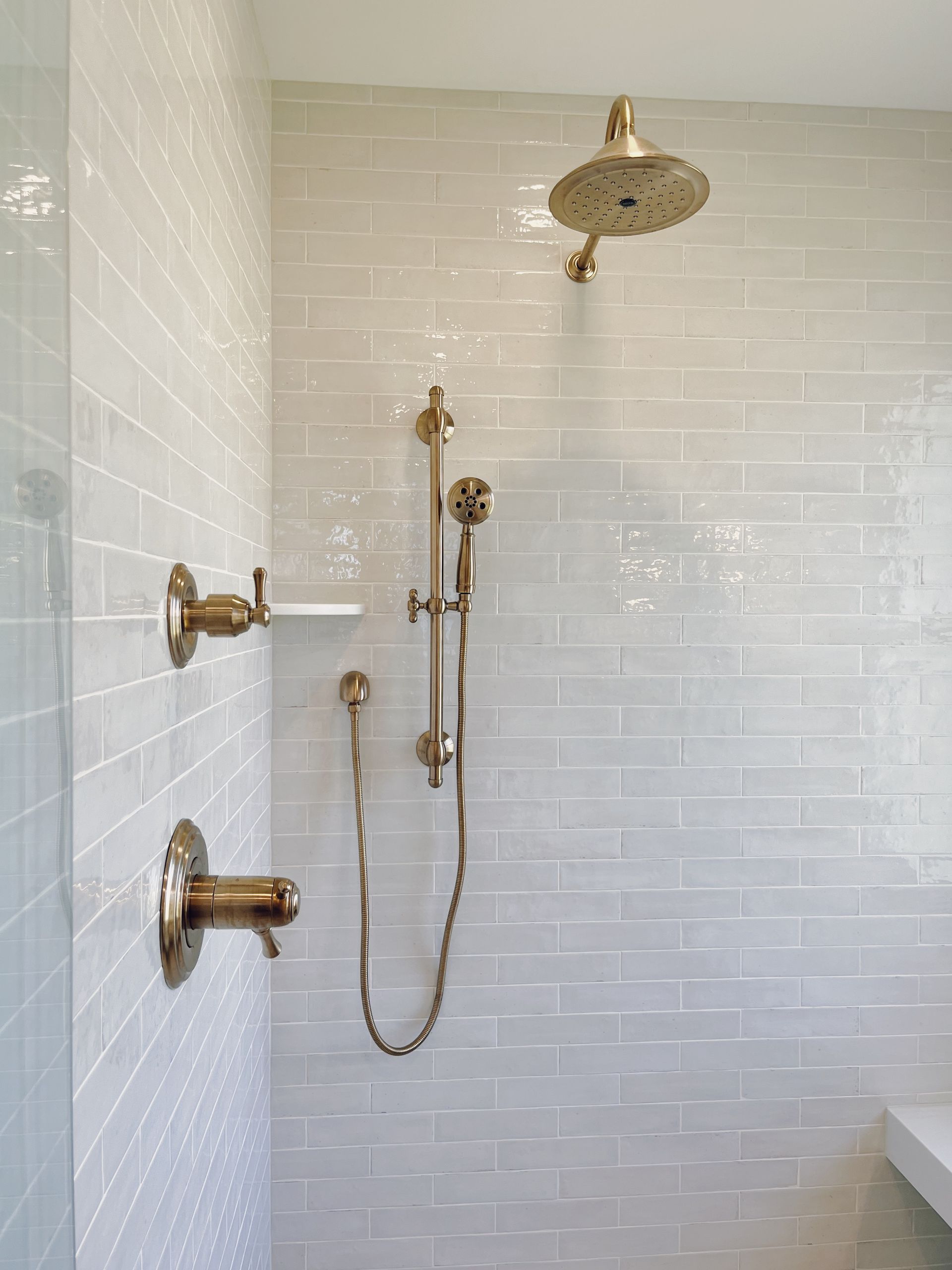 White-tiled shower with brass fixtures, handheld showerhead, and built-in bench.