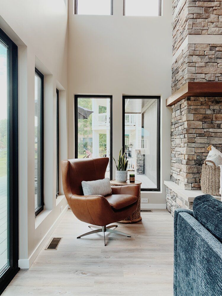 Modern living room with a brown lounge chair, tall windows, and a stone fireplace in a bright home.