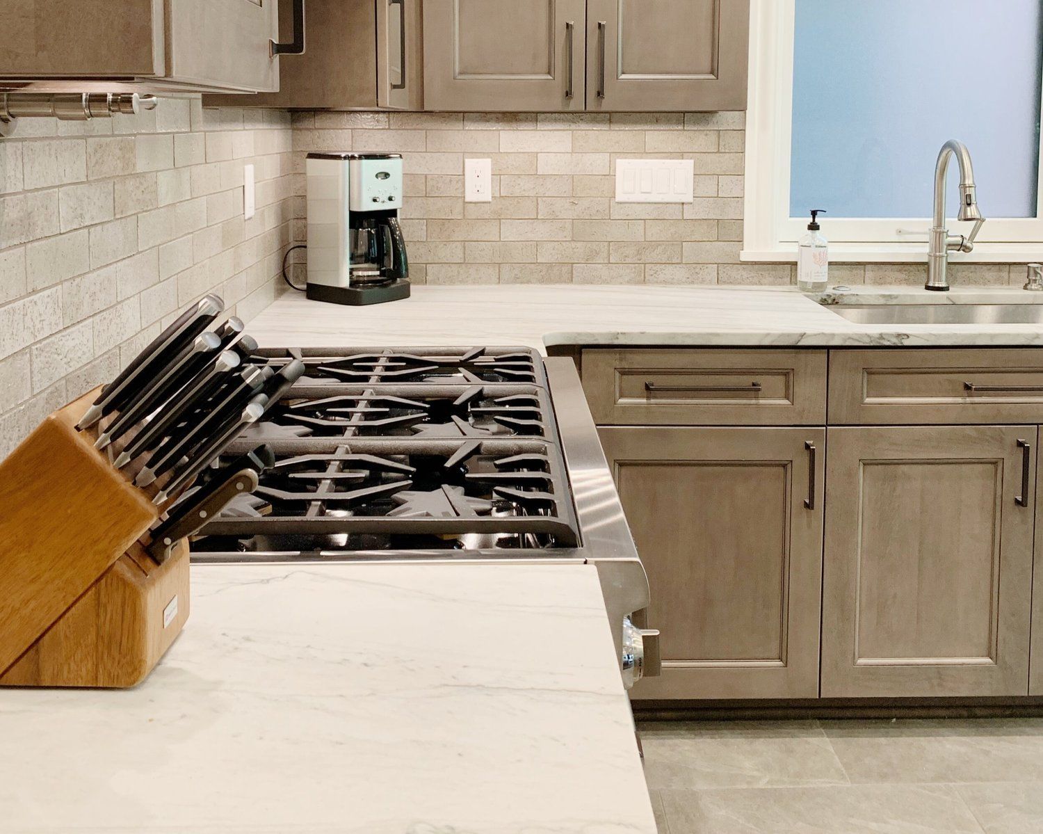 Modern kitchen with beige cabinets, white countertops, a gas stove, knife block, and sink by a blue window.