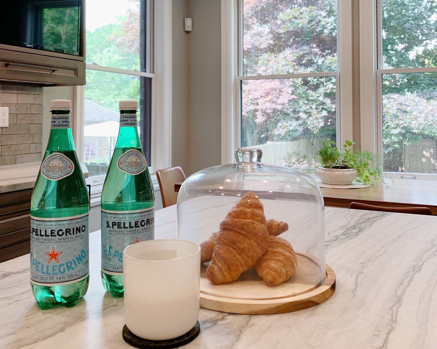 Two green sparkling water bottles beside a croissant under a glass dome on a marble kitchen counter.