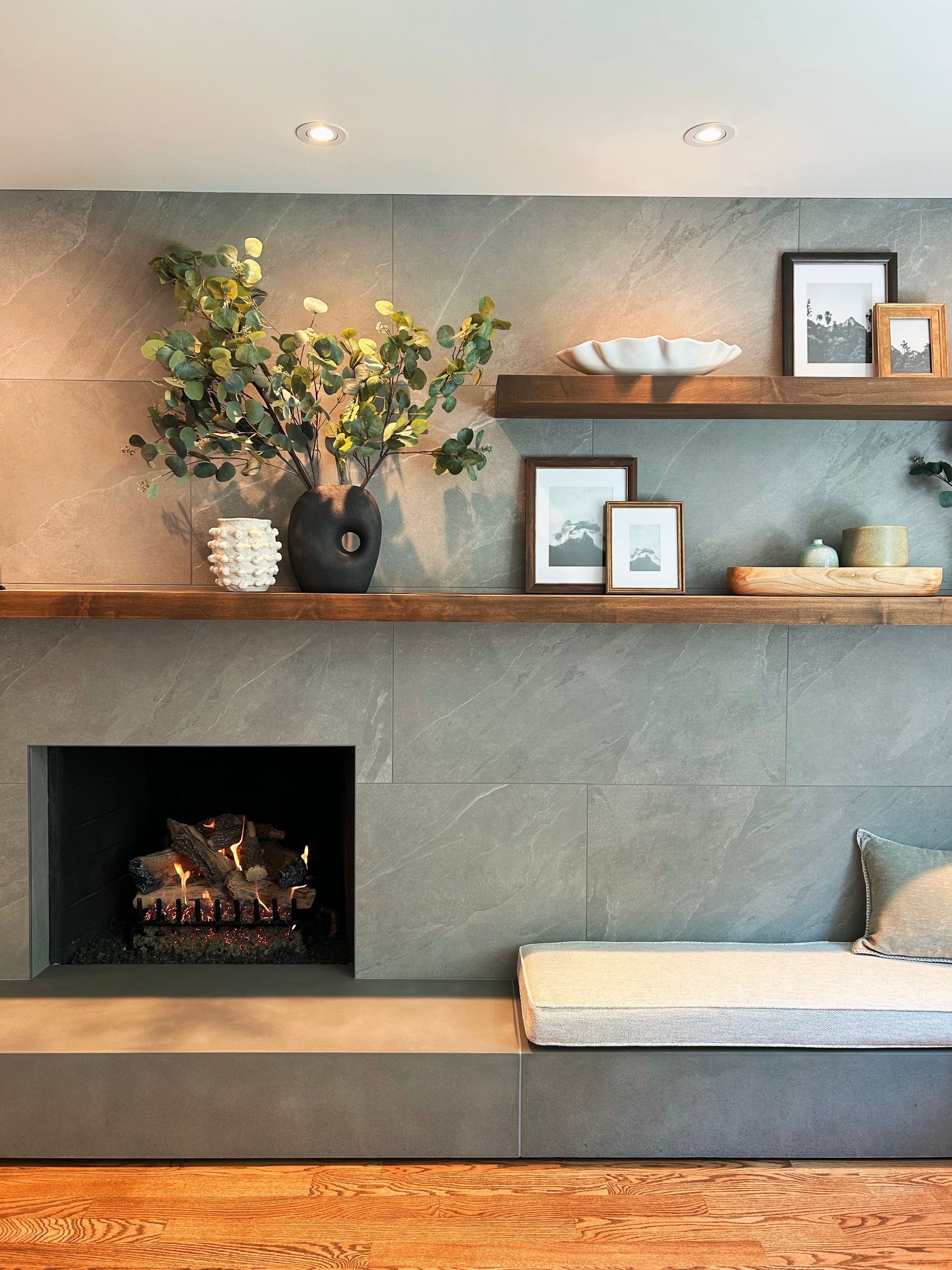 Built-in living room shelves with plants, framed art, a fireplace, and a cushioned bench in a gray modern wall.