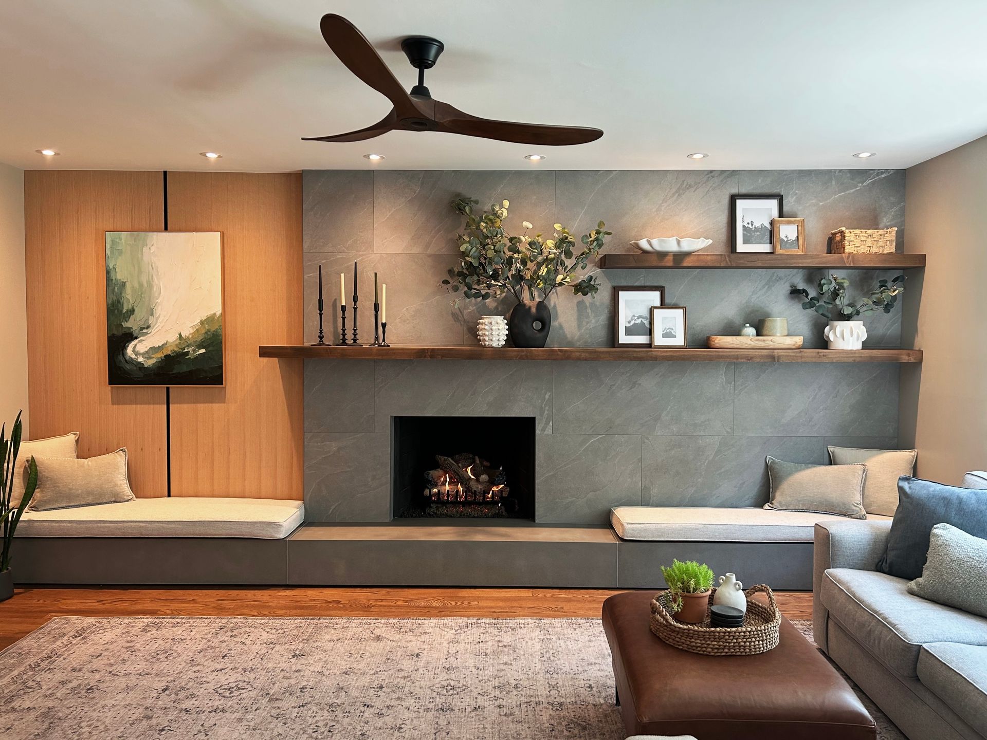 Modern living room with fireplace, gray sectional, wood shelves, and ceiling fan