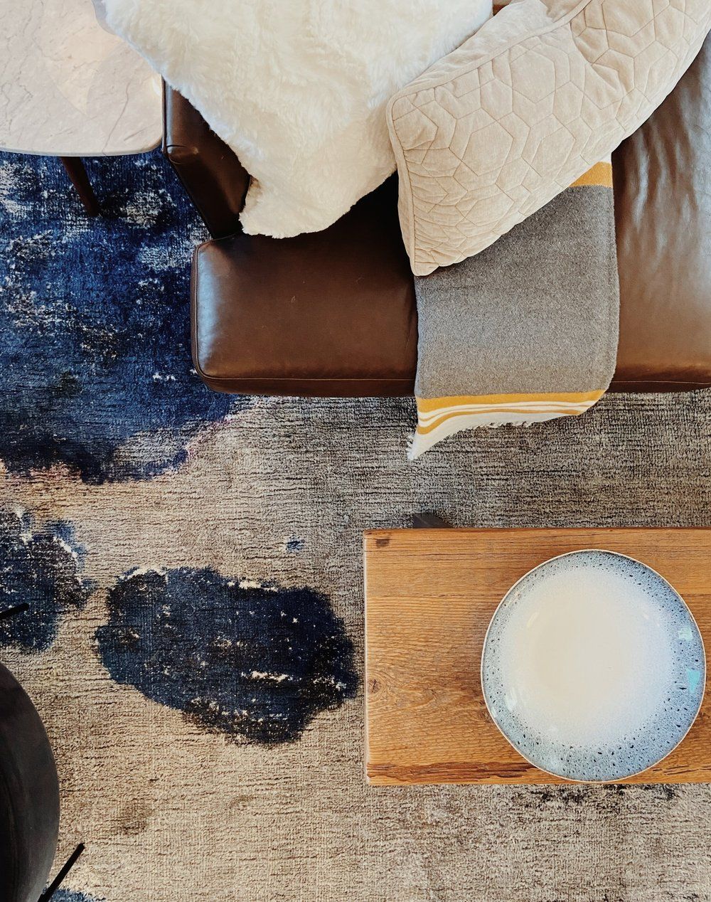 Top view of a brown sofa with cream blankets, a blue rug, and a small wooden side table holding a white bowl.
