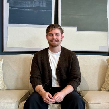Person sitting on a beige couch, smiling, in front of abstract wall art.