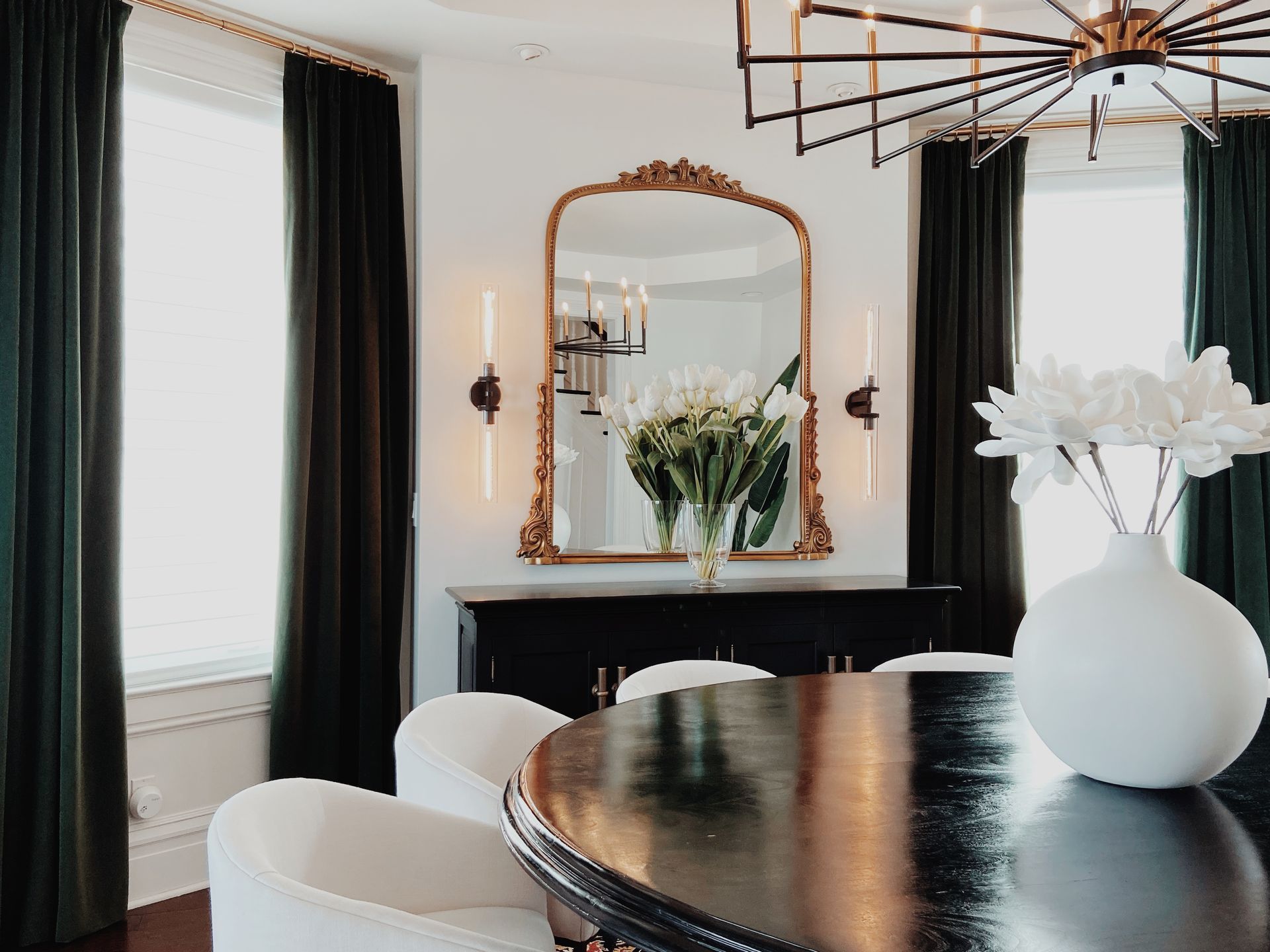 Elegant dining room with black curtains, gold mirror, black table, and white vase on a bright wall