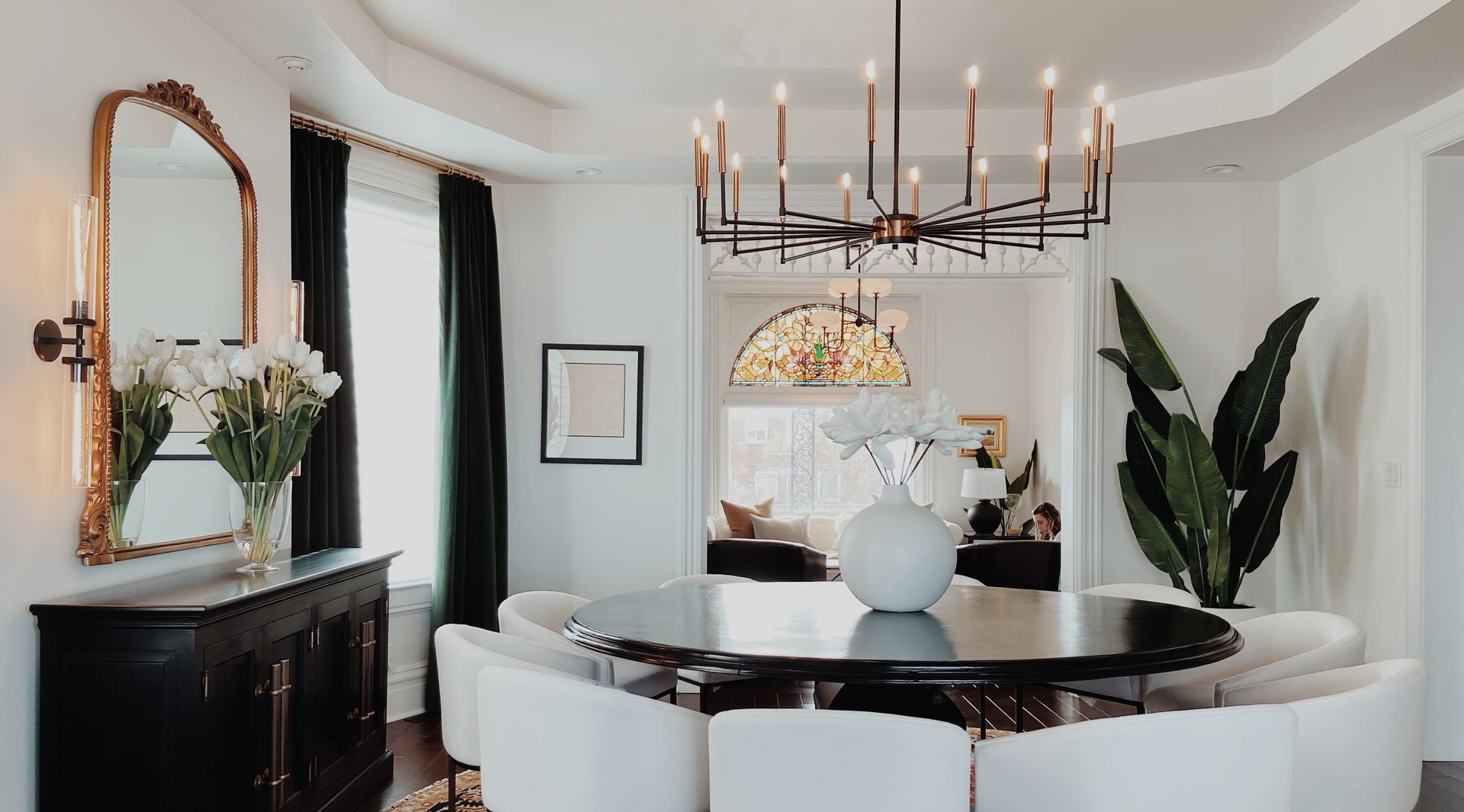Elegant dining room with round black table, white chairs, chandelier, mirror, and plants