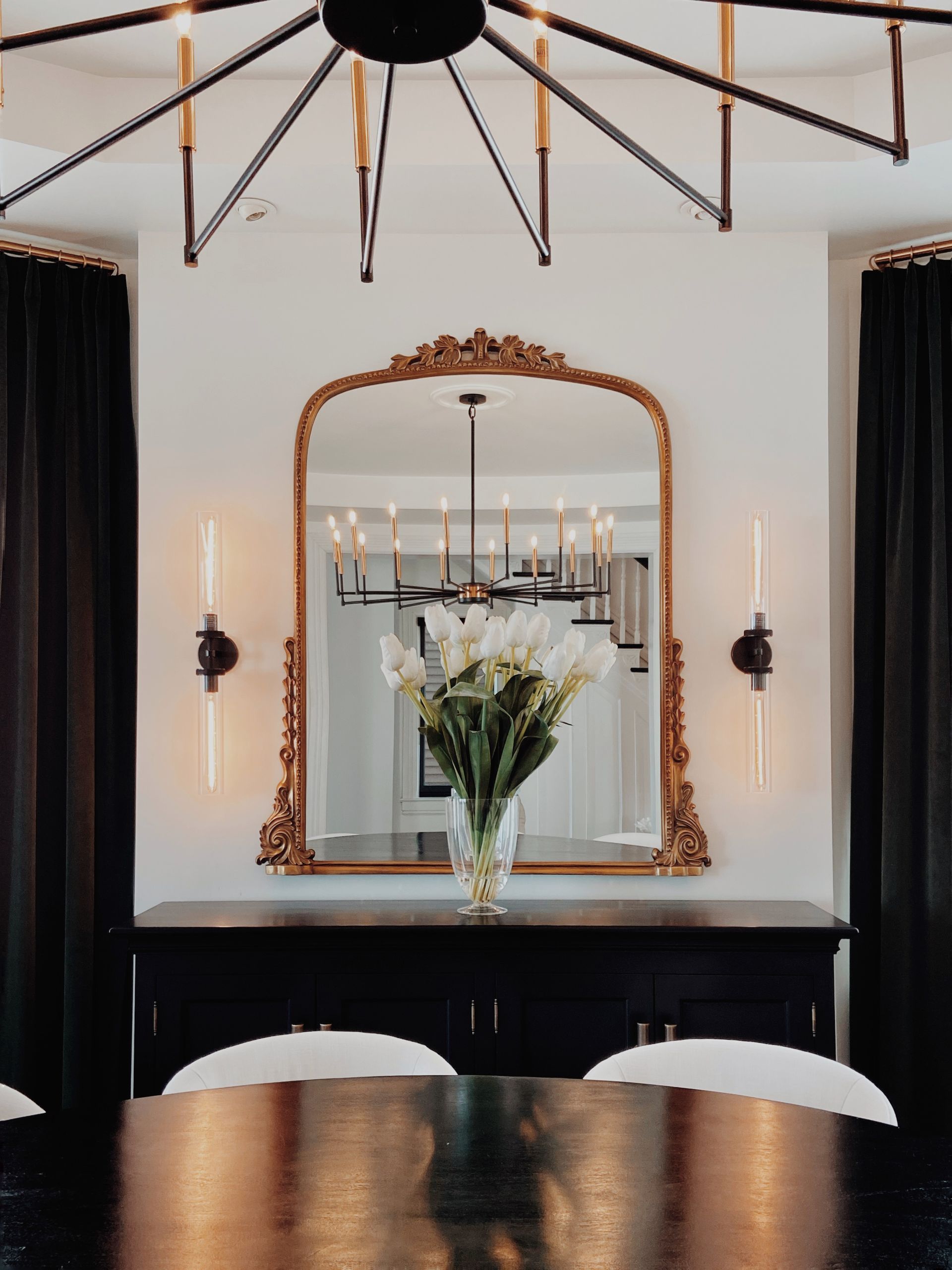Elegant dining room with ornate gold mirror, black chandelier, and table with flower vase