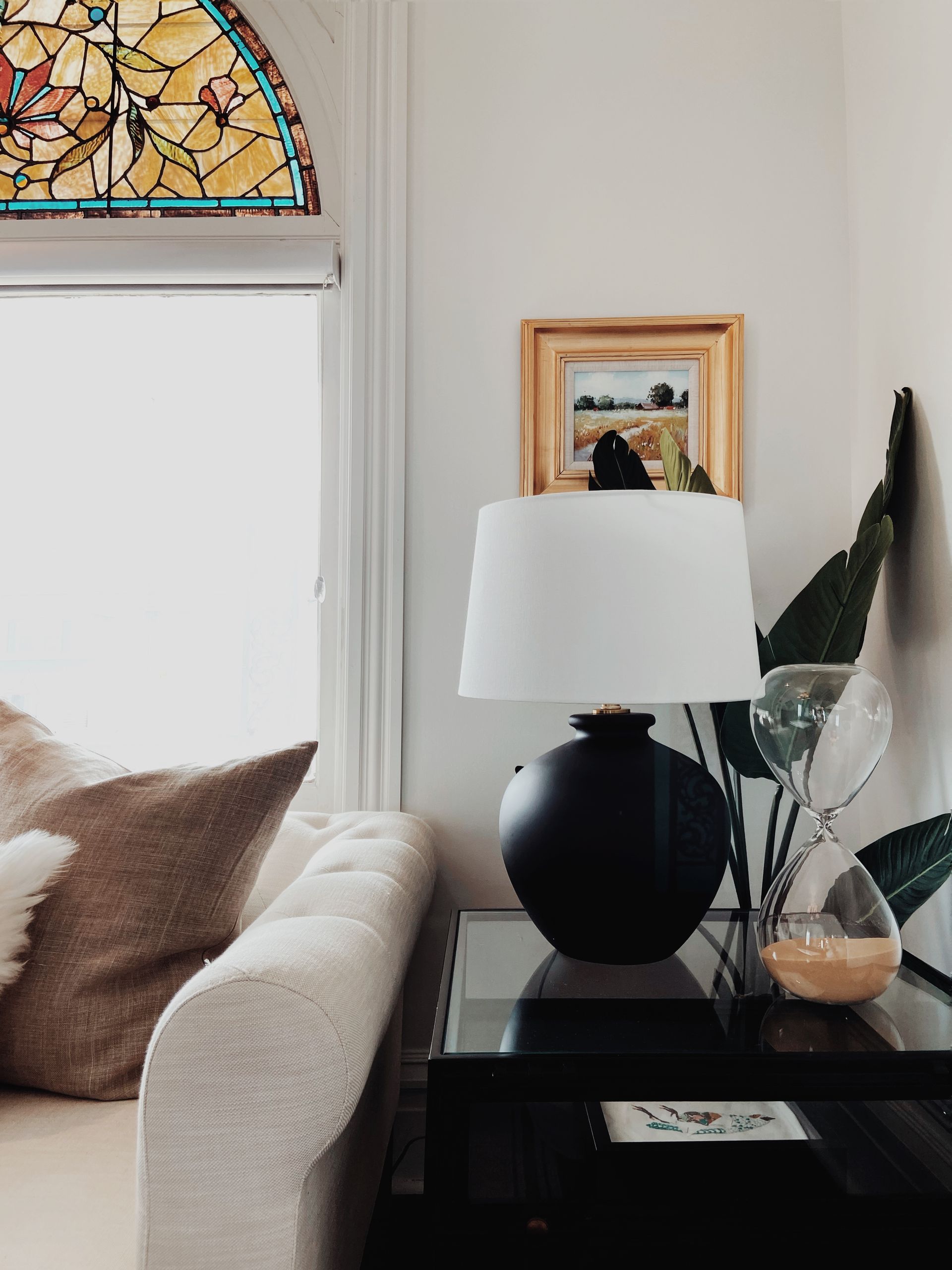 Living room corner with black lamp, side table, sofa, brown pillows, and stained glass window above