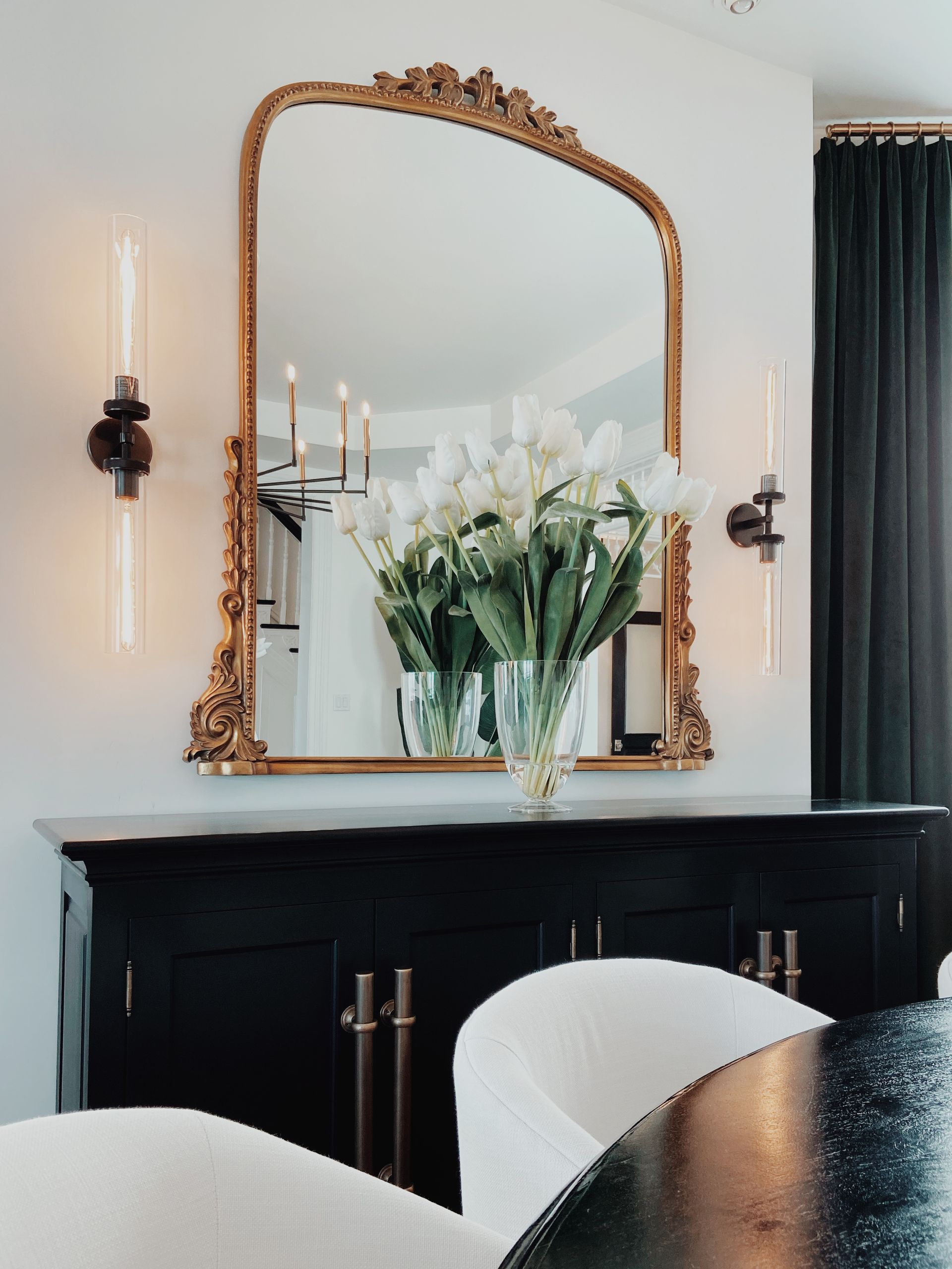 Elegant black console with gold-framed mirror, white flowers, and wall sconces in a bright room