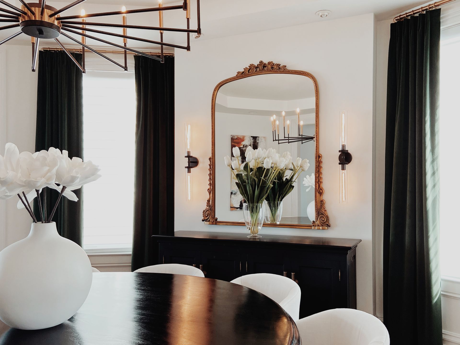 Elegant dining room with black table, white chairs, gold-framed mirror, and white flowers near tall windows
