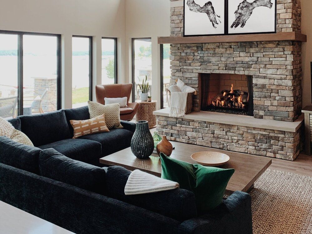 Modern living room with dark sofa, stone fireplace, and large windows overlooking a view.