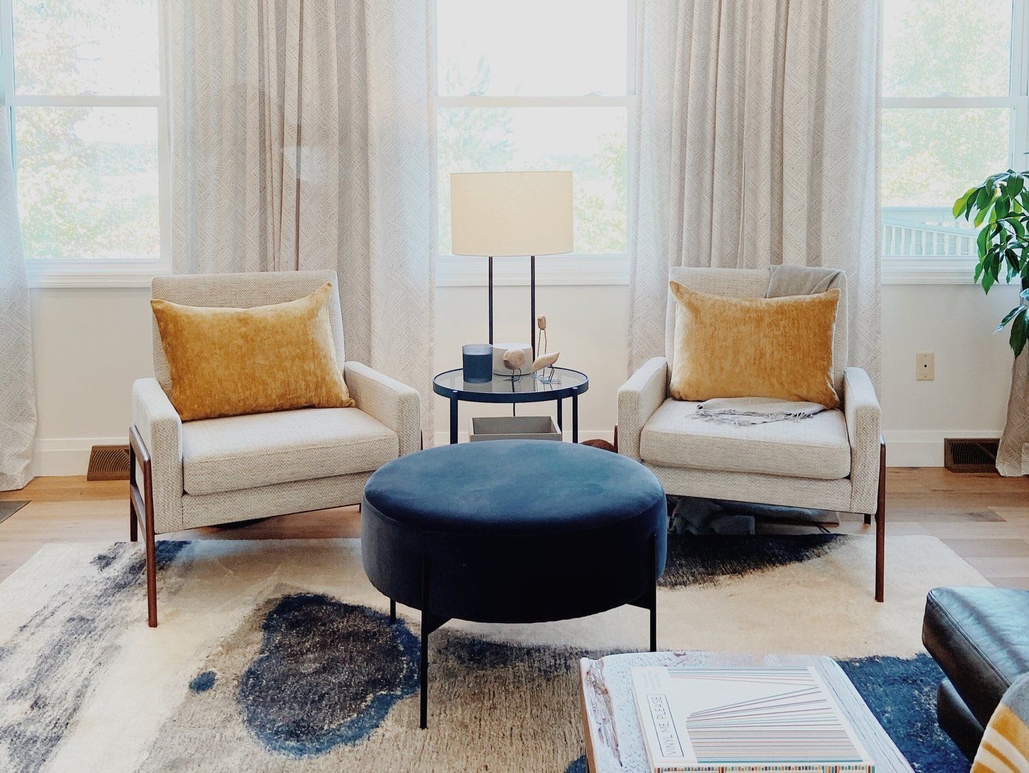 Bright living room with two cream chairs, navy ottoman, lamp, and large windows with sheer curtains