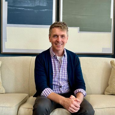Man sitting on a beige couch, smiling in a blue cardigan and checkered shirt, with abstract wall art behind him