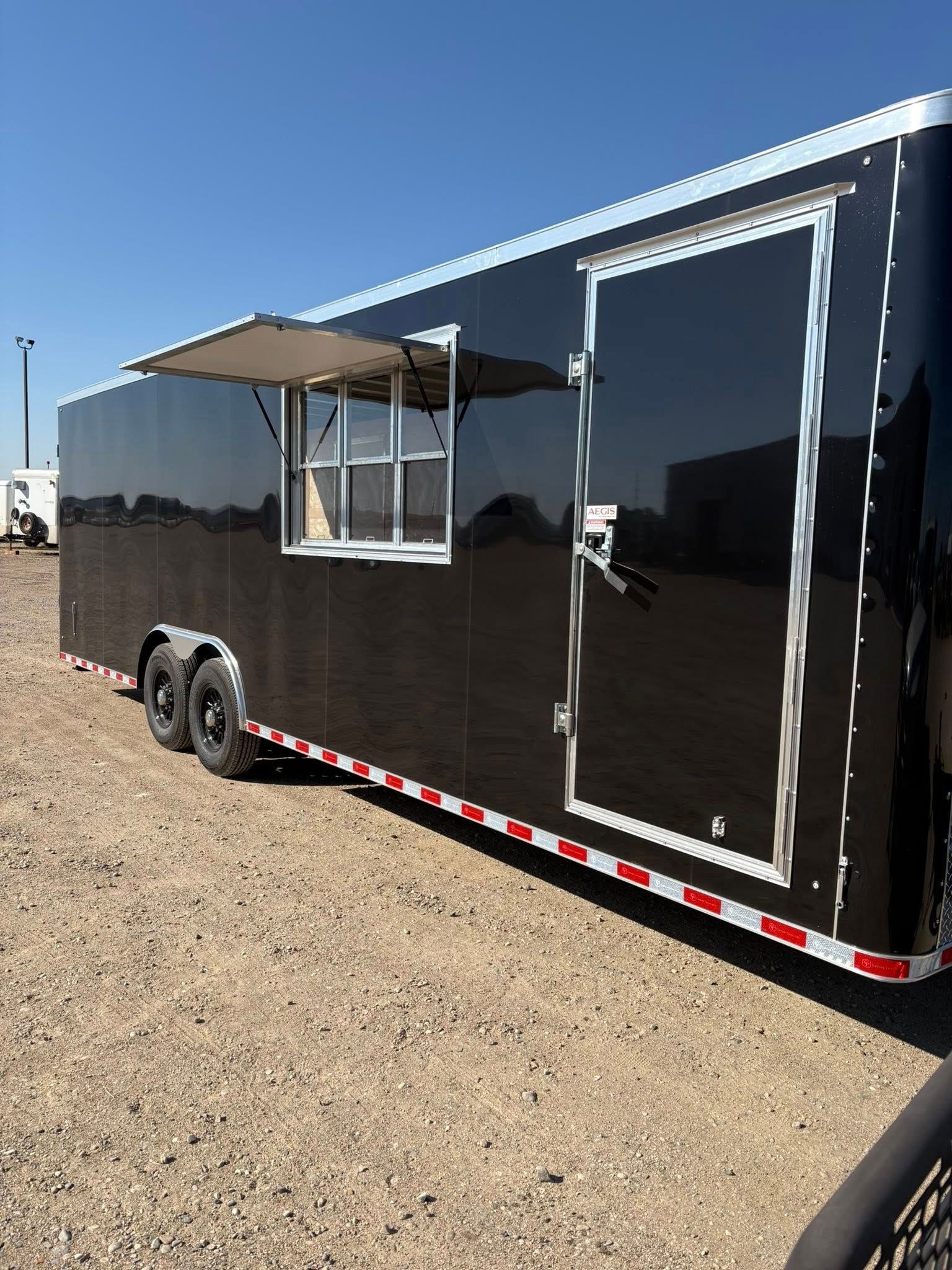 A black, dual-axle concession trailer parked outdoors on gravel with an open service window and a side entry door.