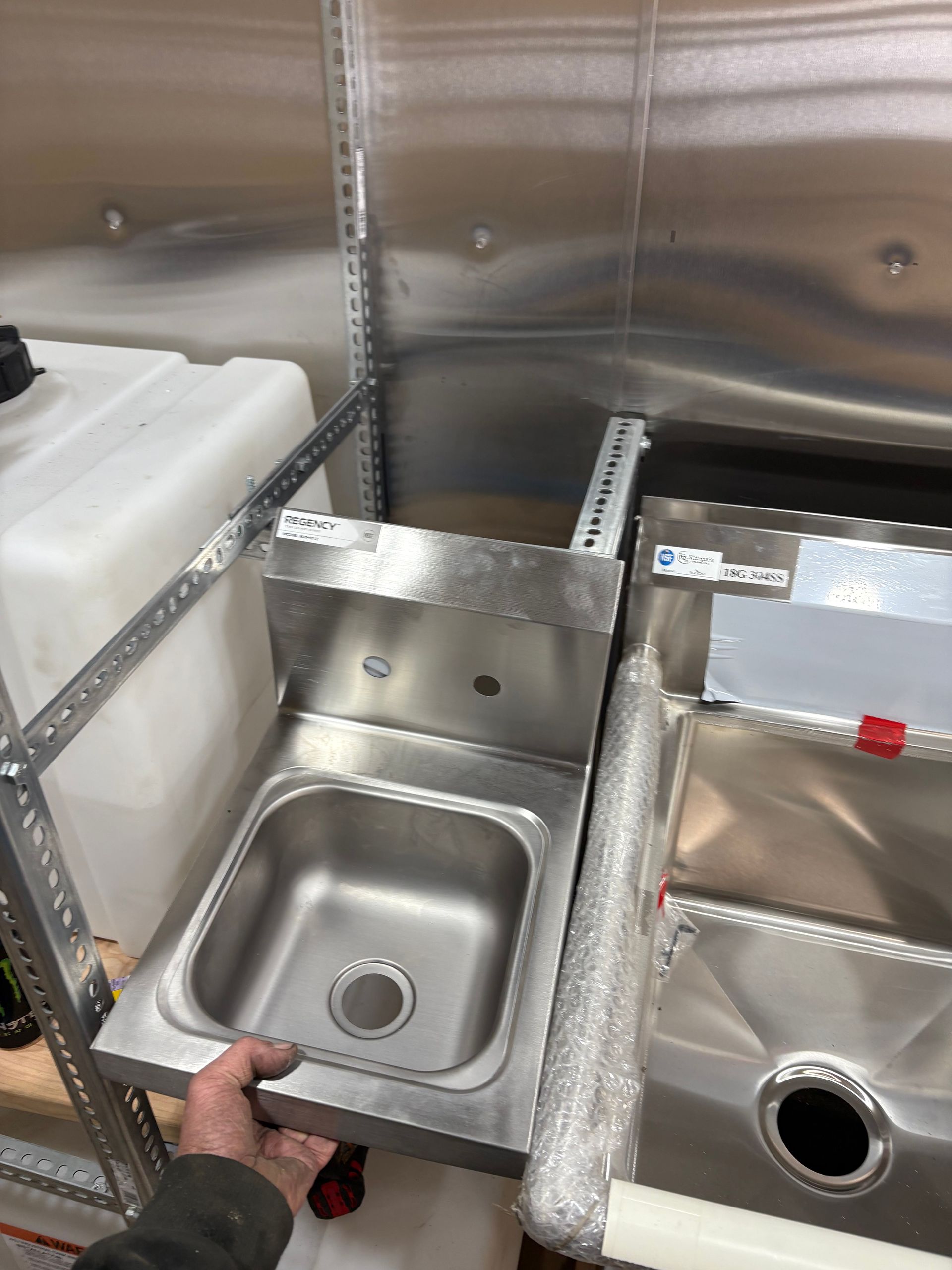 A hand holds a small stainless steel sink basin next to a larger, wrapped sink unit inside a storage or workshop area.