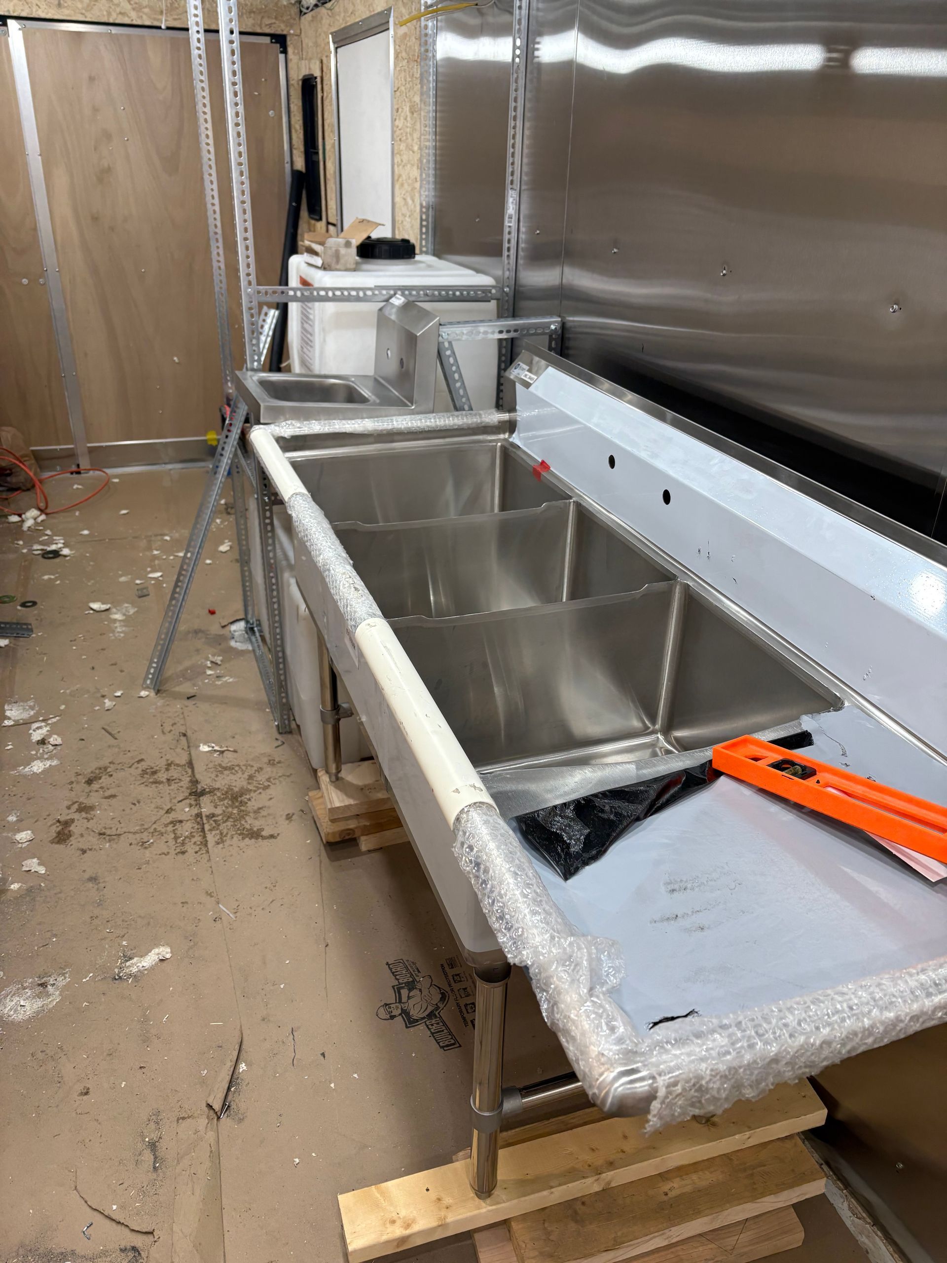 A three-compartment stainless steel commercial sink sits on wooden pallets inside an unfinished room.