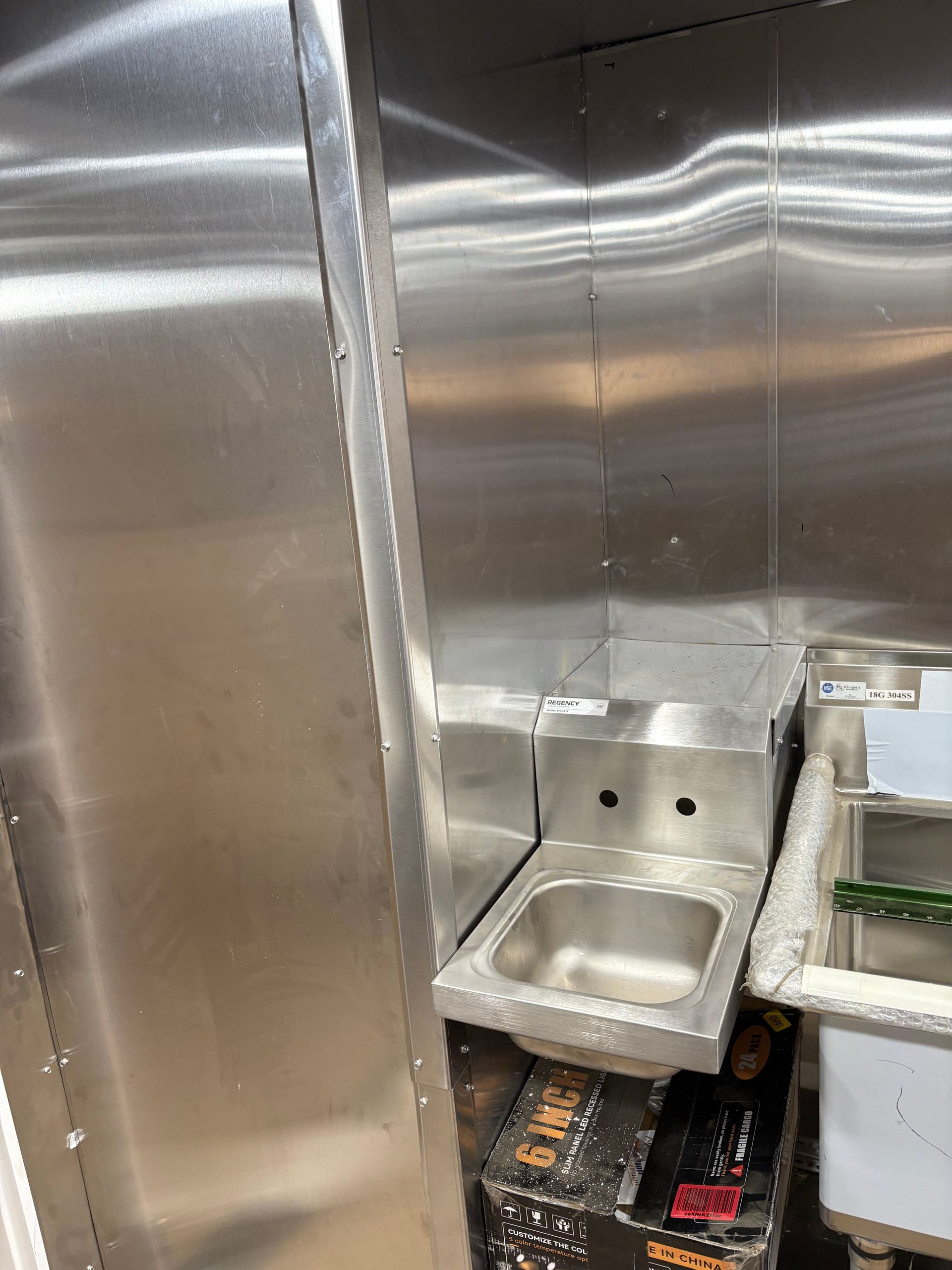 A stainless steel handwashing sink mounted on a wall in a commercial kitchen, positioned above a cardboard box.