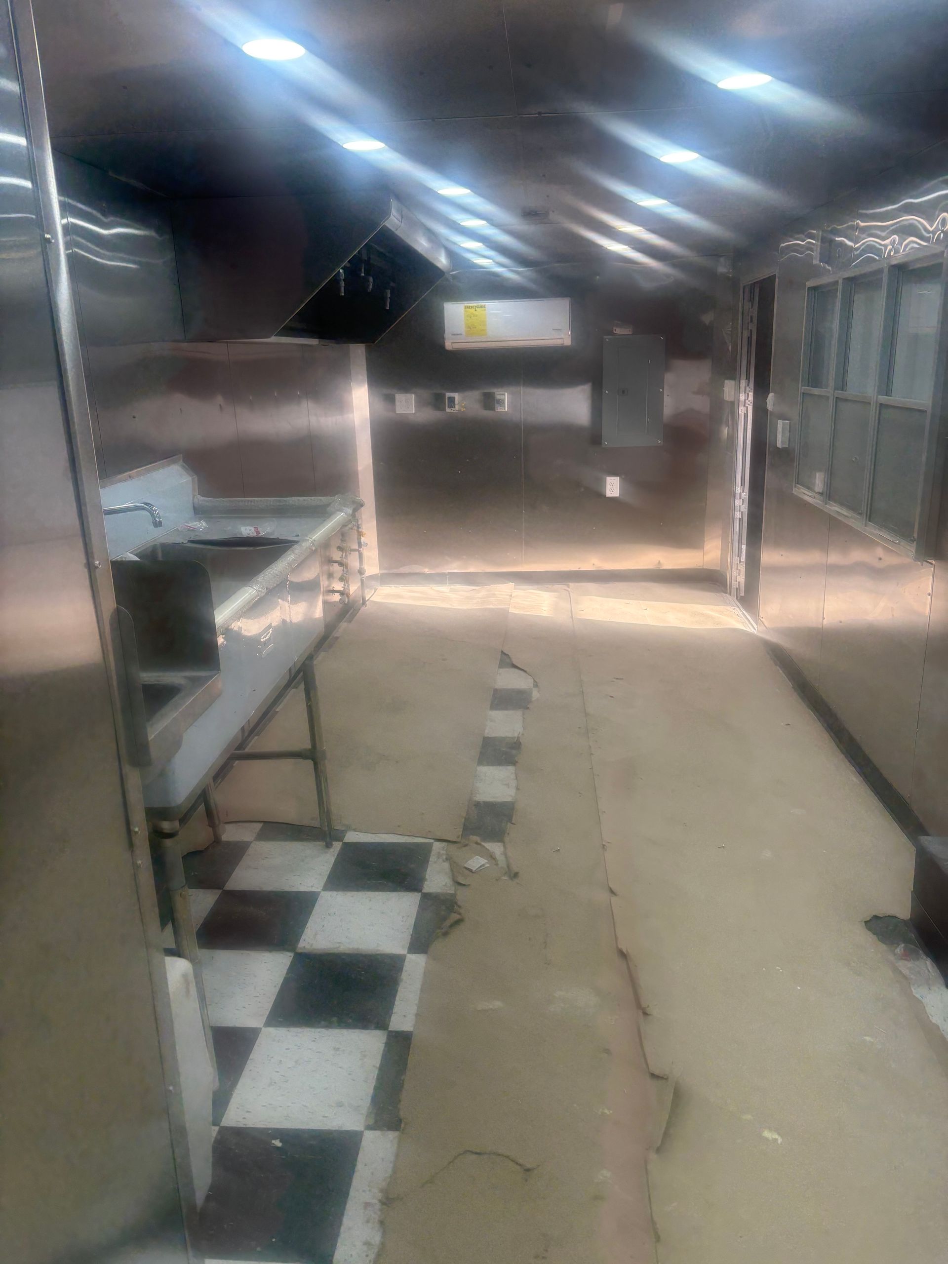 A stainless steel food prep area with a sink and a checkered floor patch in a dimly lit, narrow commercial space.