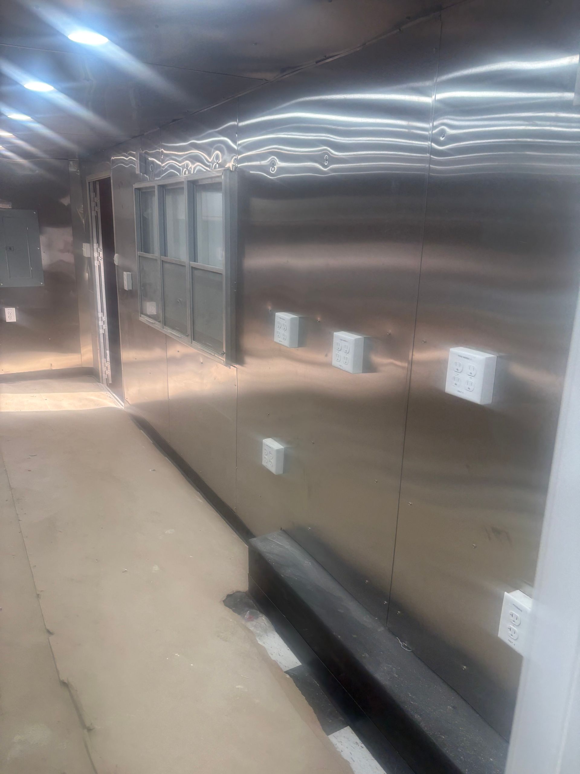 A long, metallic wall in a corridor featuring evenly spaced white electrical outlets and a series of framed windows.