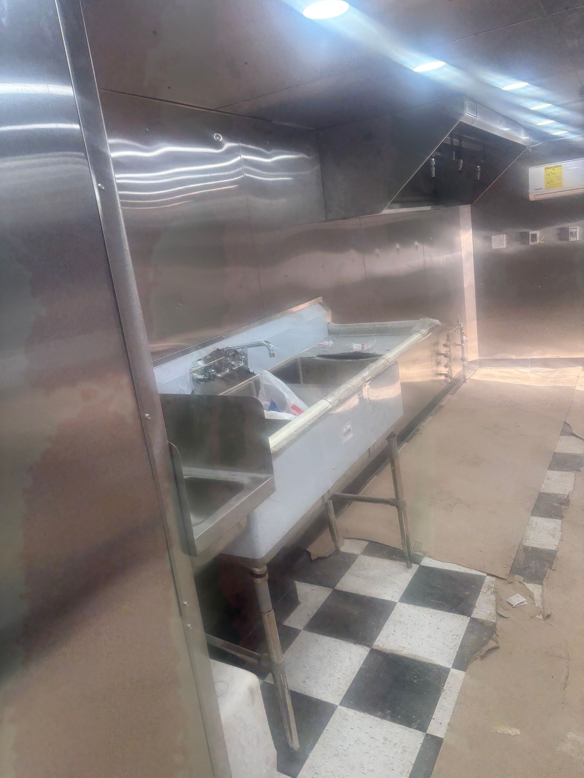 A commercial stainless steel sink with a backsplash stands on a floor with a black-and-white checkered tile pattern.