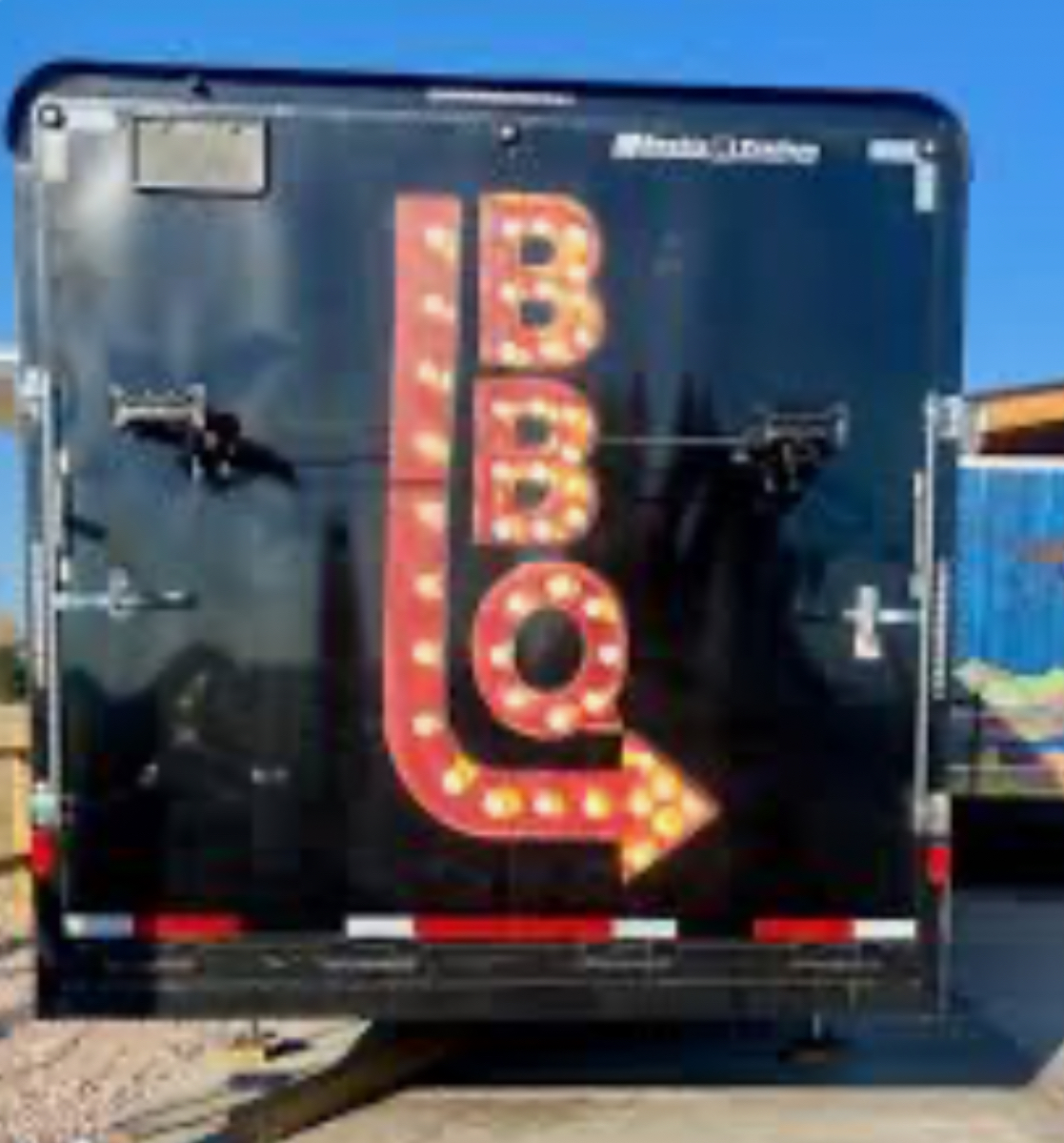The back of a black BBQ trailer features a large, illuminated sign with red, bulb-lit 