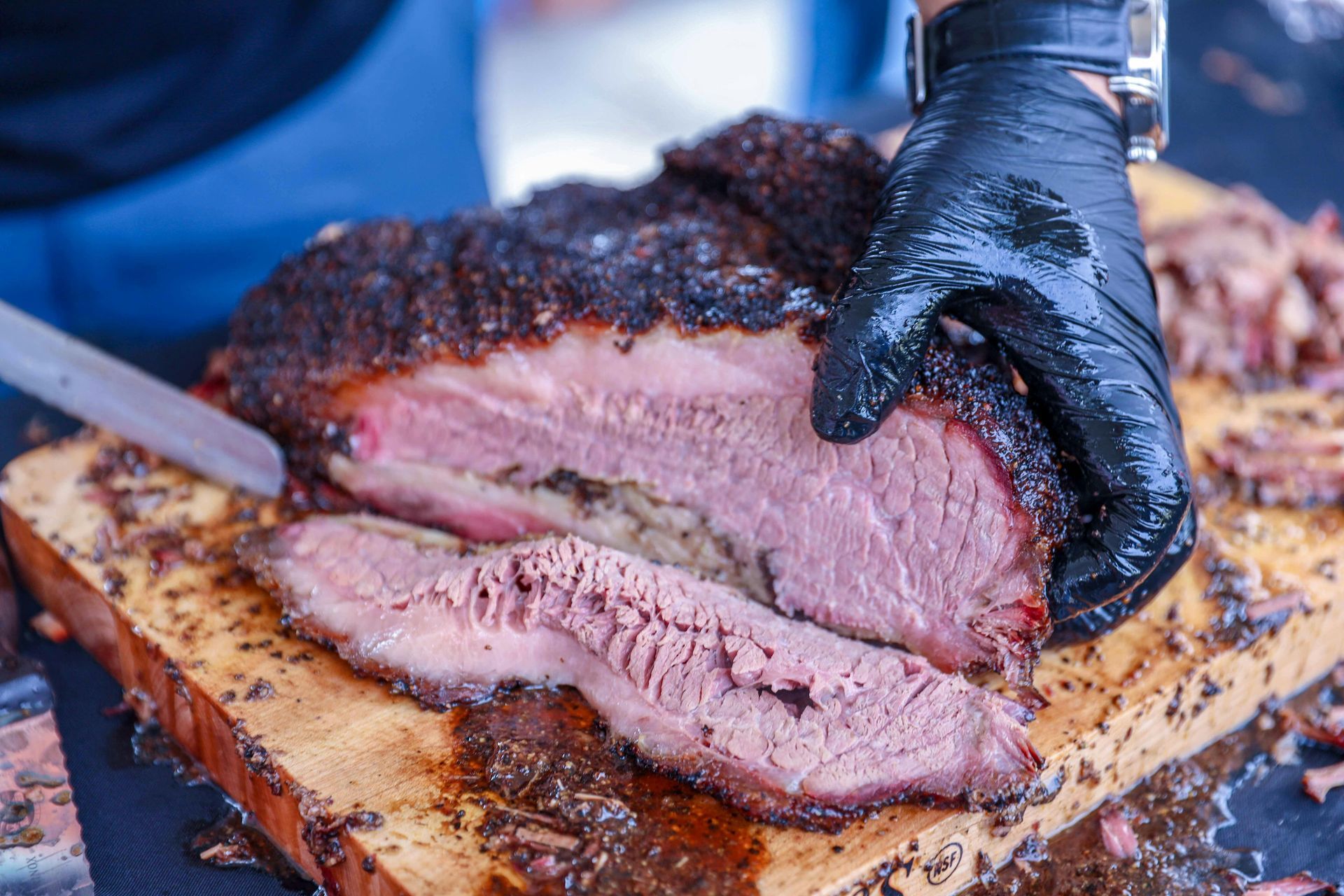A gloved hand holds a smoked, sliced beef brisket on a wooden cutting board.