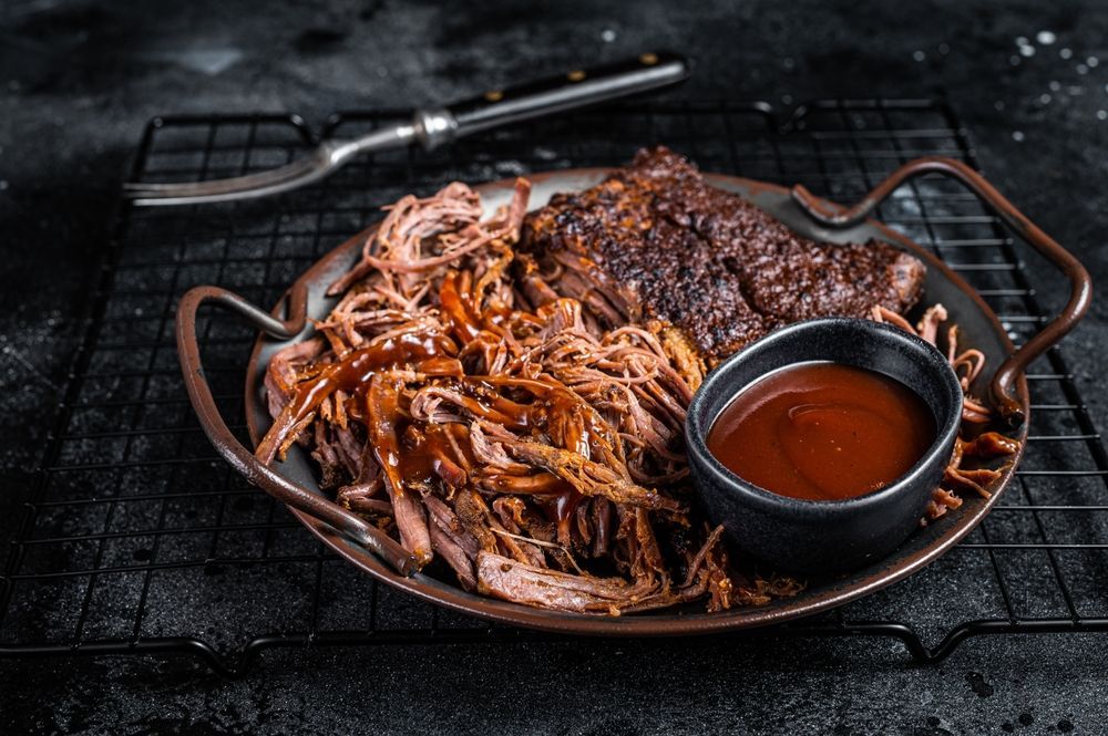 Pulled pork and a slab of smoked meat with a side of BBQ sauce on a small metal tray atop a black wire rack.