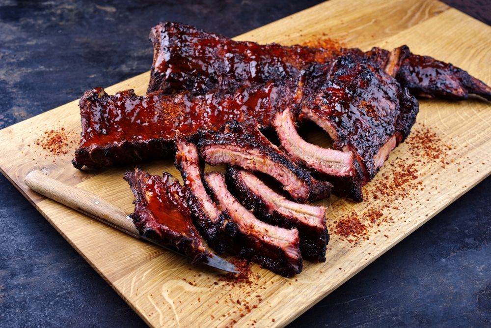 A rack of glazed, saucy barbecue pork ribs on a wooden cutting board with a knife and scattered seasoning.