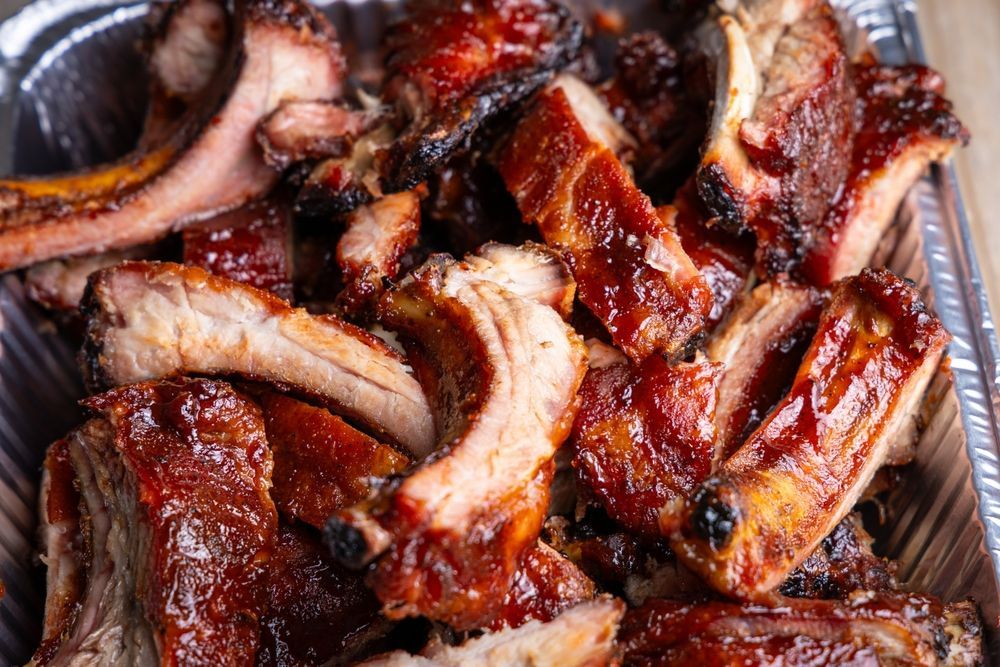 A close-up of several glazed, barbecued pork ribs arranged inside a silver aluminum catering tray.