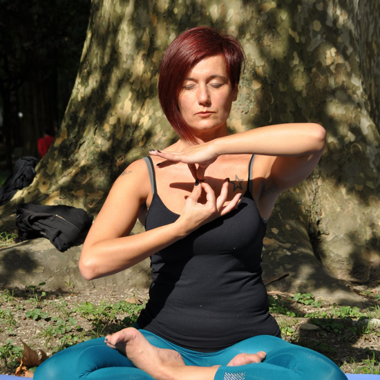Donna con canotta nera e pantaloni blu in meditazione all'aperto sotto un albero, con le mani in un mudra.