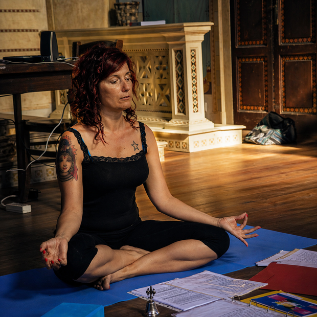 Donna con canotta nera e pantaloni blu in meditazione all'aperto sotto un albero, con le mani in un mudra.