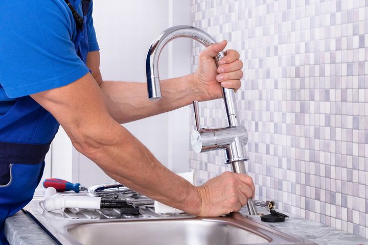 Plumber installing a chrome faucet in a kitchen sink; tools visible on counter.