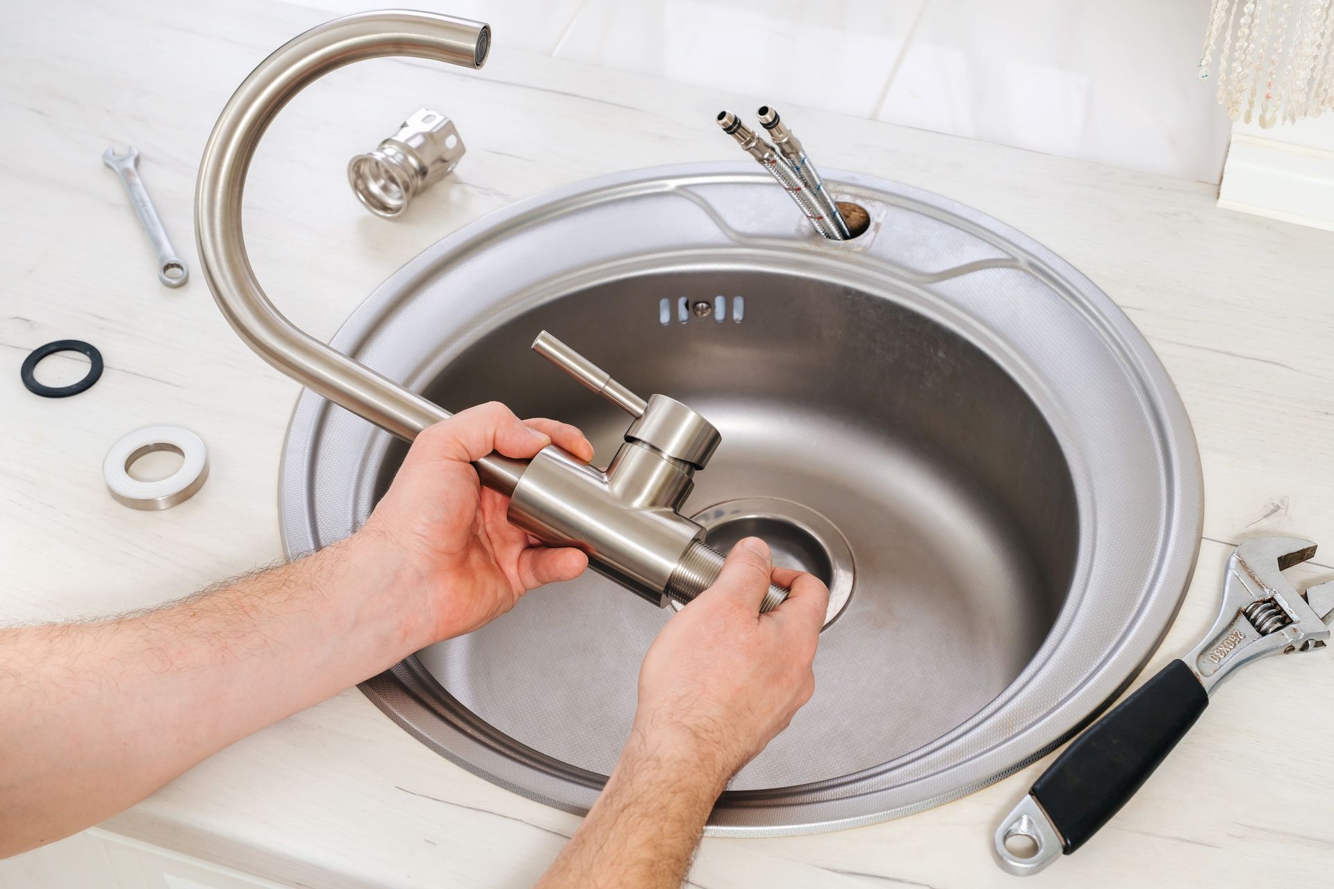 Hands installing a new kitchen faucet in a stainless steel sink, with tools and parts nearby.