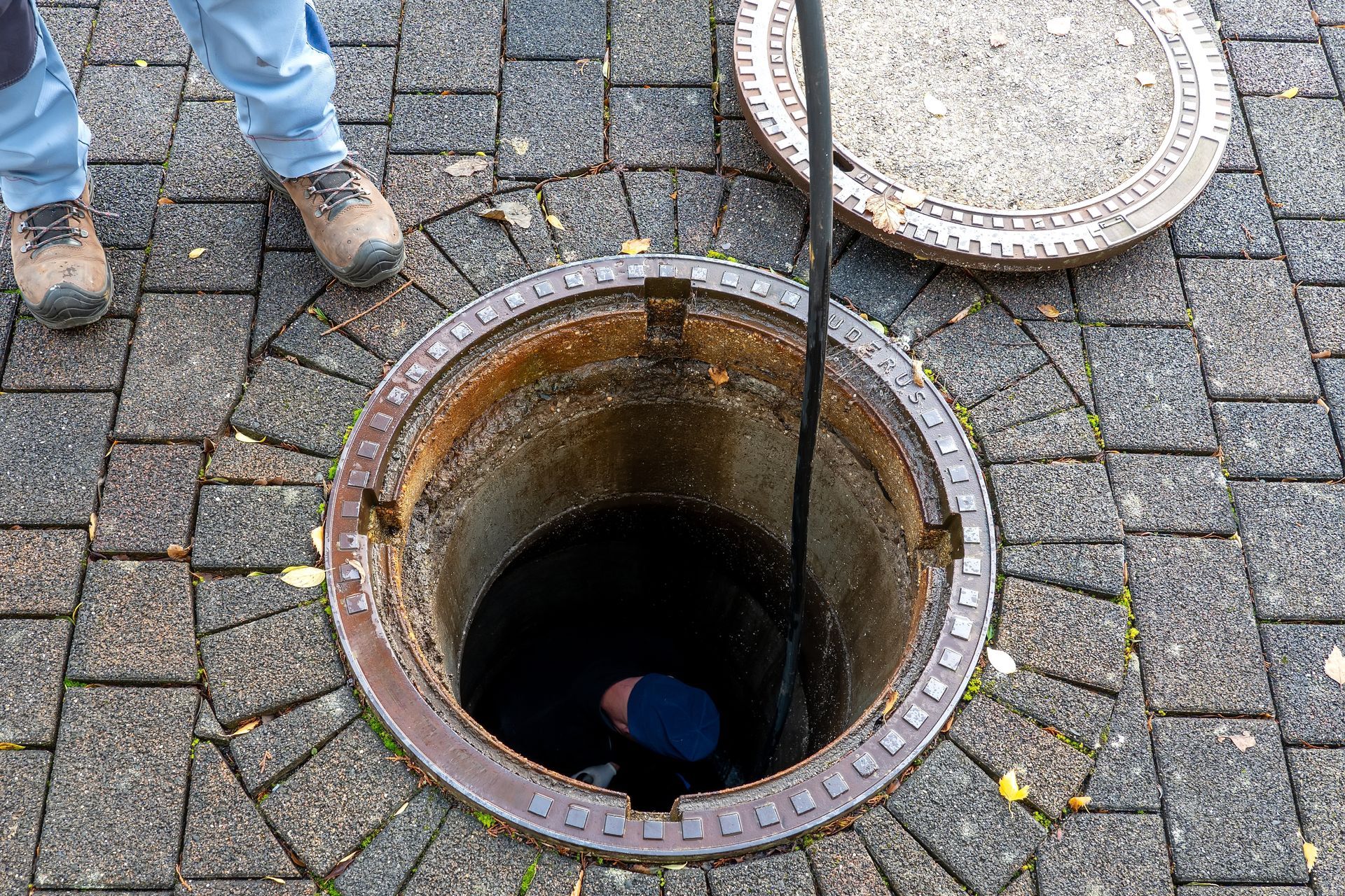 Manhole open on a brick-paved surface; a person inside with a rope; lid nearby and legs visible above.