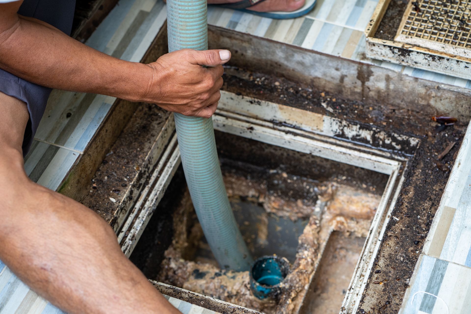 Person using a hose to clean a dirty drain in a tiled setting.