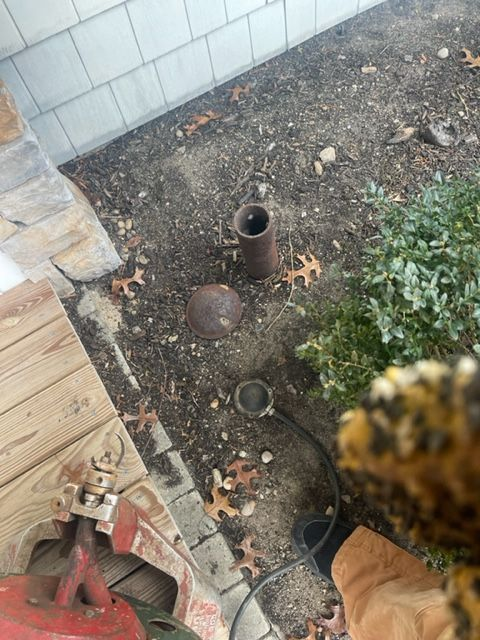 Rusty pipes and a cap in a dirt patch beside a building. Some leaves and greenery present.